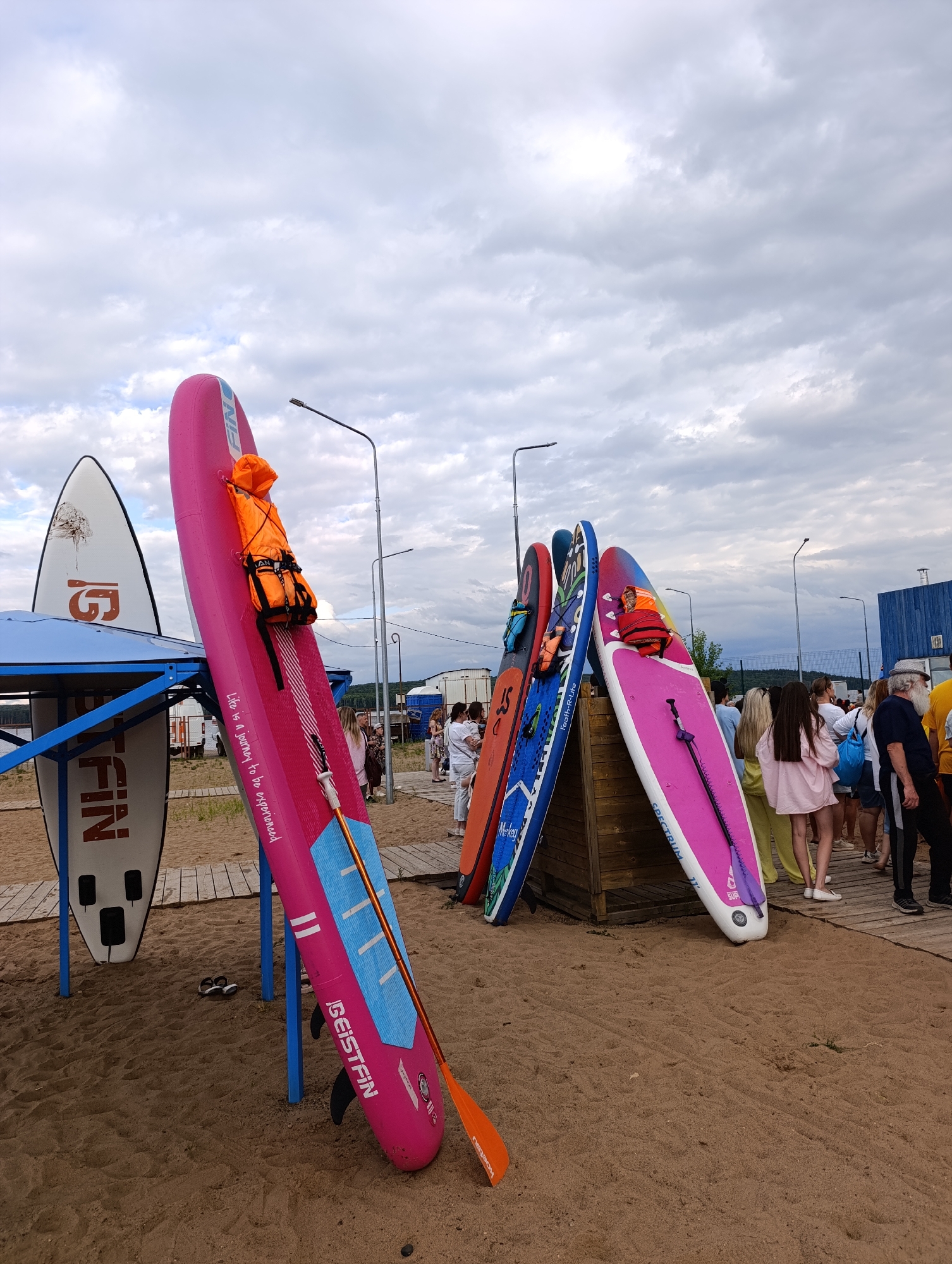 This is a full-shot, eye-level, outdoor photograph taken on a sandy beach with a wooden boardwalk in Chaikovsky, Russia, under a cloudy sky. The scene features several stand-up paddleboards (SUPs) leaning against a wooden structure and a blue metal frame. The foreground is dominated by sand.
In the center of the image, a bright pink SUP is prominently displayed, leaning at an angle. It has a life vest with an orange hue attached to its upper section, and text reading "Life is a journey to be experienced" is visible vertically on its side, along with the word "GEISTFIN". A paddle with an orange blade and a purple shaft rests on the pink board.
To the left of the pink SUP, a white SUP with a graphic and text that reads "FIN" is supported by a blue metal frame. Behind these, other SUPs are visible, including a blue one with colorful graphics and an orange one.
In the background, a crowd of people are gathered on the boardwalk, some appearing to be waiting or socializing. There are streetlights and some distant structures, including what appears to be a blue industrial building. The overall impression is a recreational area during a cloudy day.