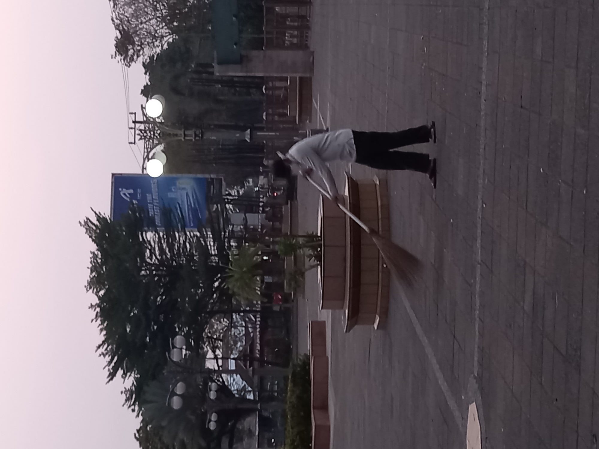 A person wearing a light-colored long-sleeved shirt, dark pants, and sandals is sweeping a paved outdoor area with a broom. The area appears to be a plaza or walkway with patterned brick paving. In the background, there are trees, streetlights, a building with a traditional roof, and a large advertisement billboard. The scene is captured during what appears to be early morning or late evening, with soft, diffused light. The overall environment suggests an urban setting, possibly a public park or square within Samarinda, Indonesia, given the context of the request.