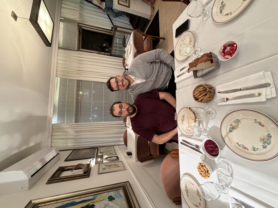 Two men wearing glasses pose at a dinner table. A table is set with plates, silverware, glasses, and small bowls of various foods, including what appears to be horseradish, nuts, and bread. Behind them are windows with curtains and other tables and chairs. Hanging lights are visible in the ceiling. Artwork hangs on the left wall.
