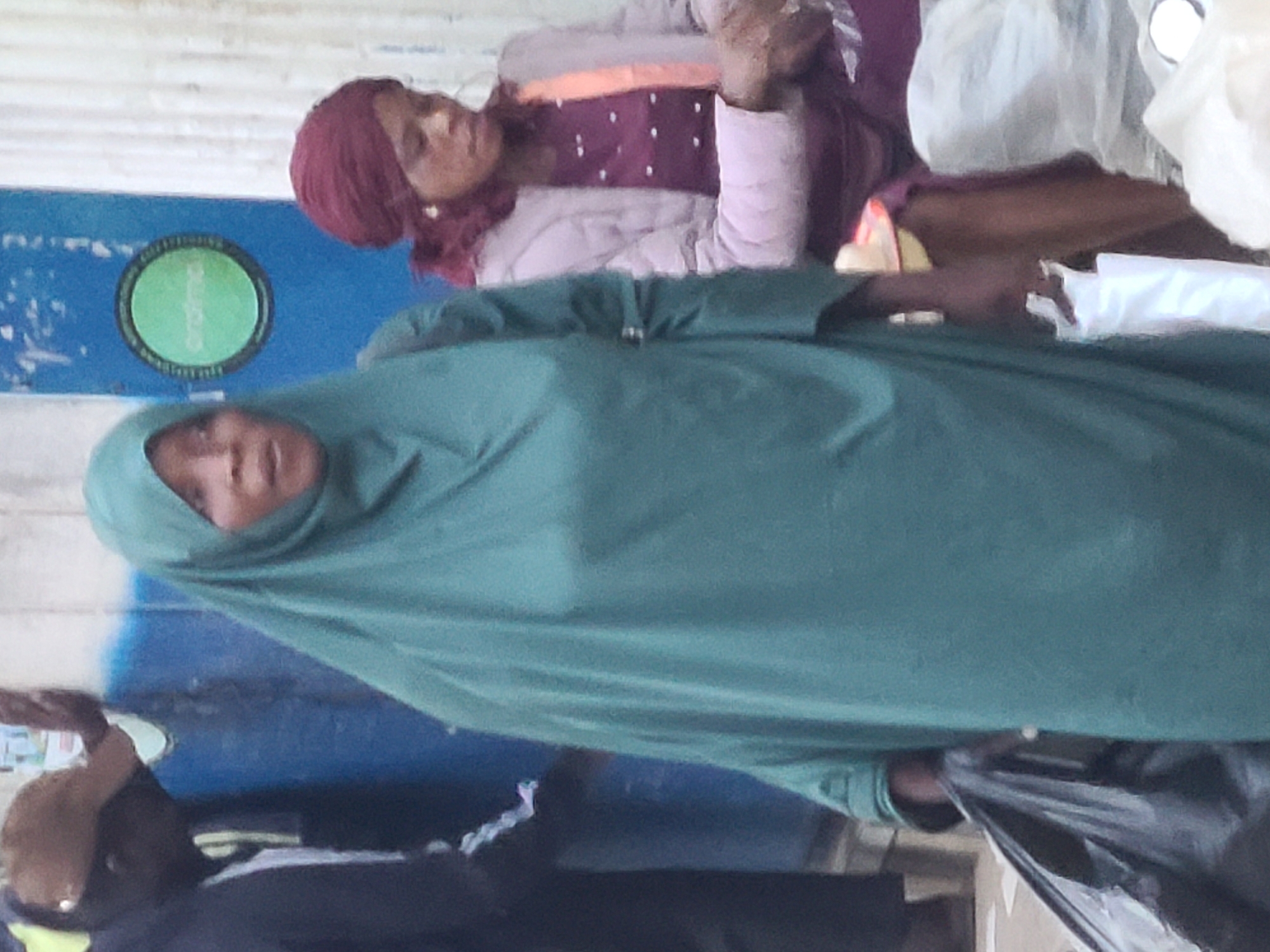 The image depicts a street scene, likely in Jos, Nigeria. In the foreground, a woman wearing a teal hijab and a matching long dress is holding a black plastic bag in her left hand. She is facing slightly to her right and appears to be speaking or looking in that direction. Behind her and to her right, another woman is visible, wearing a maroon headscarf and a light-colored, possibly padded jacket over a purple, polka-dotted dress. She is looking down and to her right, her hands are obscured. To the far left, a man in a dark jacket and a brown baseball cap is partially visible, leaning against a blue wall. The background features a blue painted wall with a circular green logo. The ground appears to be paved. The overall impression is of a busy street or market environment.