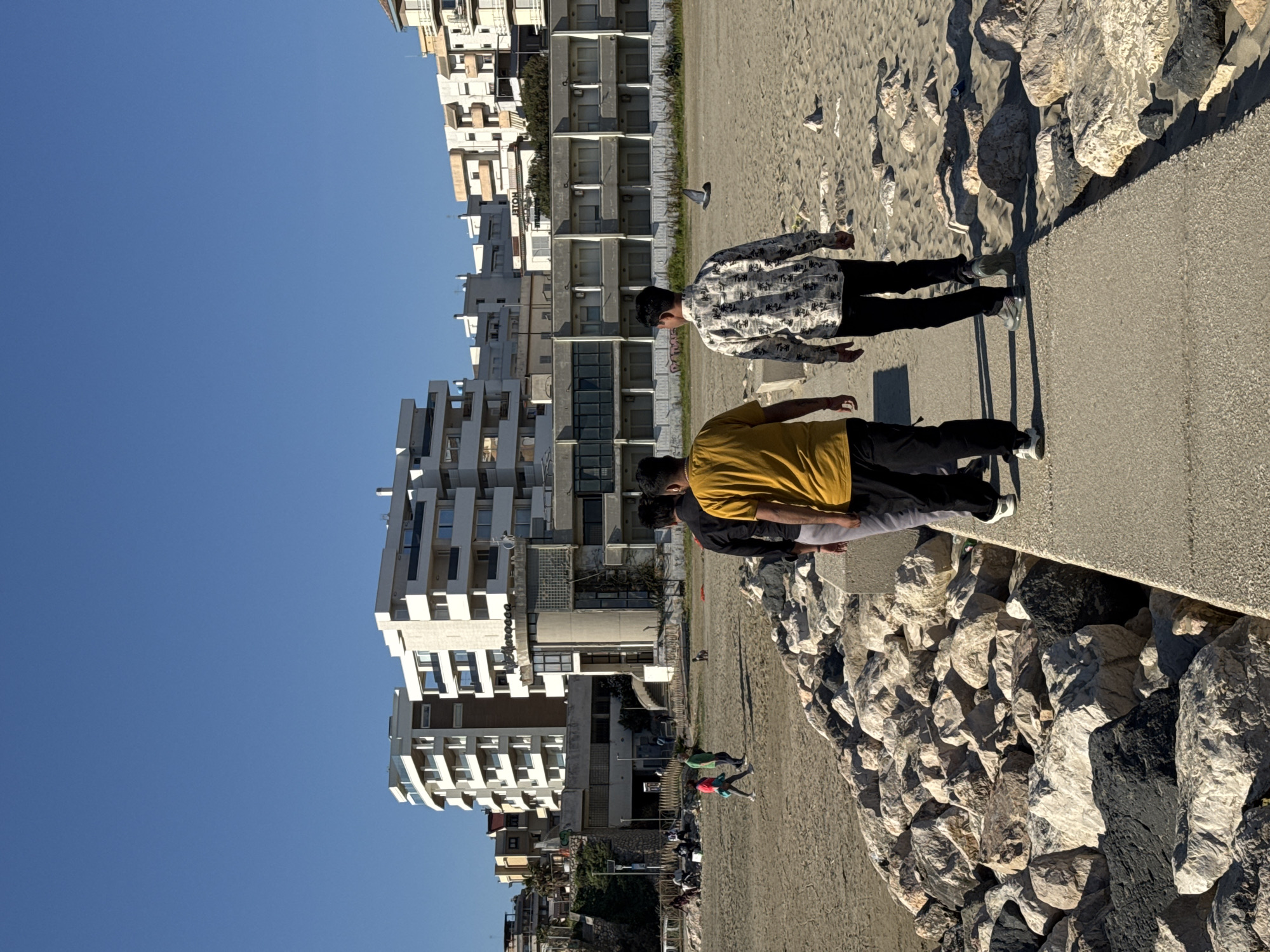 The image captures a sunny coastal scene in Nettuno, Italy, featuring a group of people by the sea with urban buildings in the background. The perspective is from a concrete promenade or path that runs alongside a rocky embankment.
In the immediate foreground, three men are standing on this concrete path, facing towards the sandy beach and the sea beyond. The man closest to the viewer wears a yellow t-shirt and dark pants, while the man in front of him has a black and white patterned long-sleeved shirt with dark pants and light sneakers. Another person with a dark shirt is partially visible between them. Their shadows are long and distinct on the path, suggesting either morning or late afternoon sunlight.
To the left of the concrete path, there is a substantial rocky barrier or sea wall, composed of various sizes of light and dark grey rocks. Beyond this barrier lies a wide, sandy beach. Several birds, possibly seagulls or pigeons, are visible scattered across the sand. Further back on the beach, more people can be seen; one individual in a bright green shirt and another in a red shirt appear to be walking or playing.
Dominating the background on the left side of the frame are numerous multi-story buildings, likely apartment complexes or hotels, lining the beachfront. These structures exhibit modern architectural styles with multiple windows and balconies, some appearing white and others in darker tones. On one of the buildings further back, the word "HOTEL" is clearly visible vertically.
The sky above is a clear, bright blue, indicating fair weather. The overall impression is one of a vibrant, sun-drenched urban beach environment.