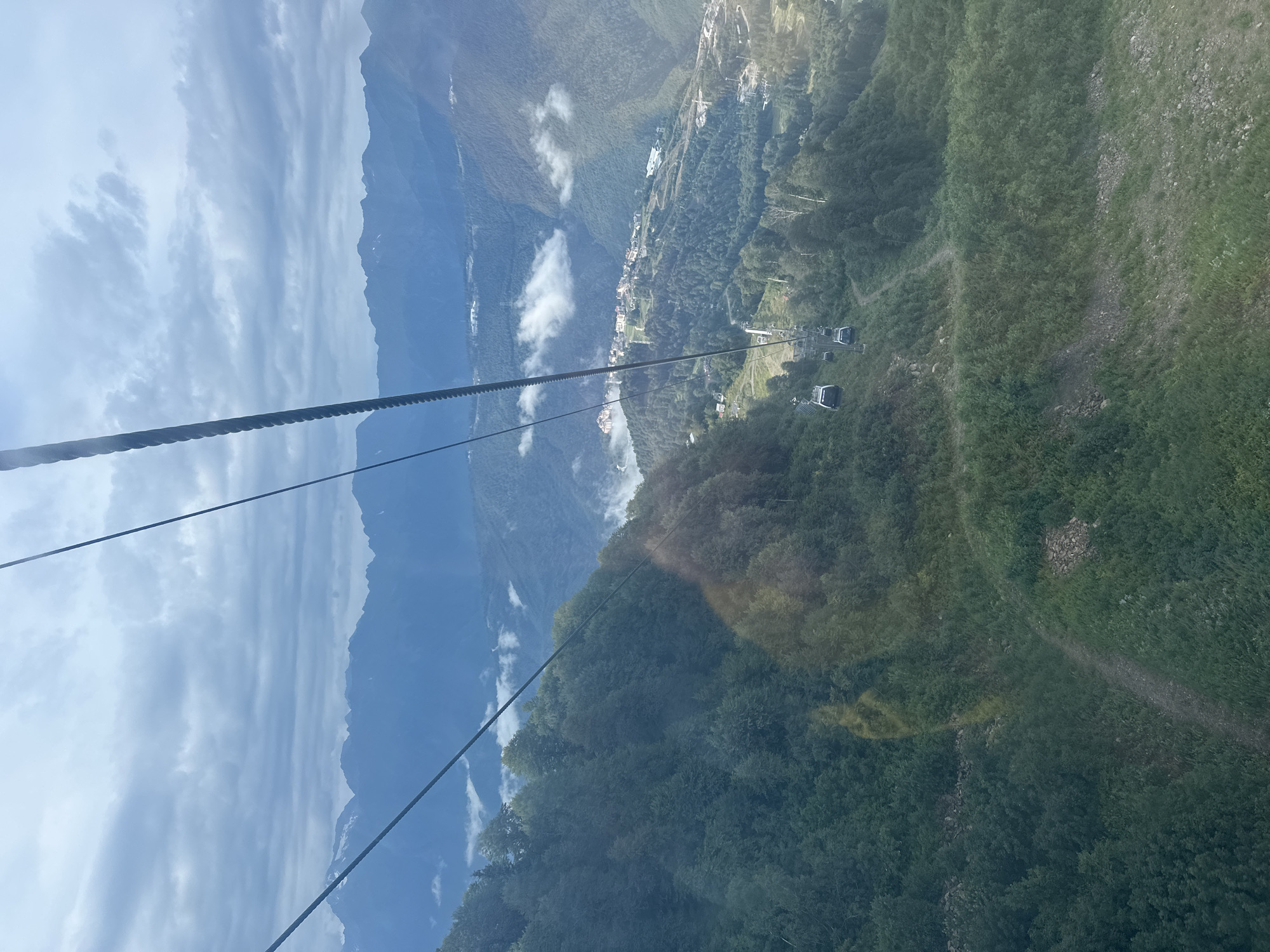 The image captures a panoramic view from a cable car descending a lush green mountainside in Estosadok, Russia. The cable car system is clearly visible, with multiple cabins suspended from thick cables. Below, the mountainside is densely covered with trees, interspersed with trails and patches of grass. In the mid-ground, a village or resort area is nestled among the mountains, with buildings and infrastructure visible. Further in the distance, a range of majestic mountains, partially shrouded in clouds, forms the backdrop. The sky is overcast with dramatic, textured clouds, and rays of sunlight break through in some areas. The overall scene conveys a sense of natural beauty and outdoor recreation.