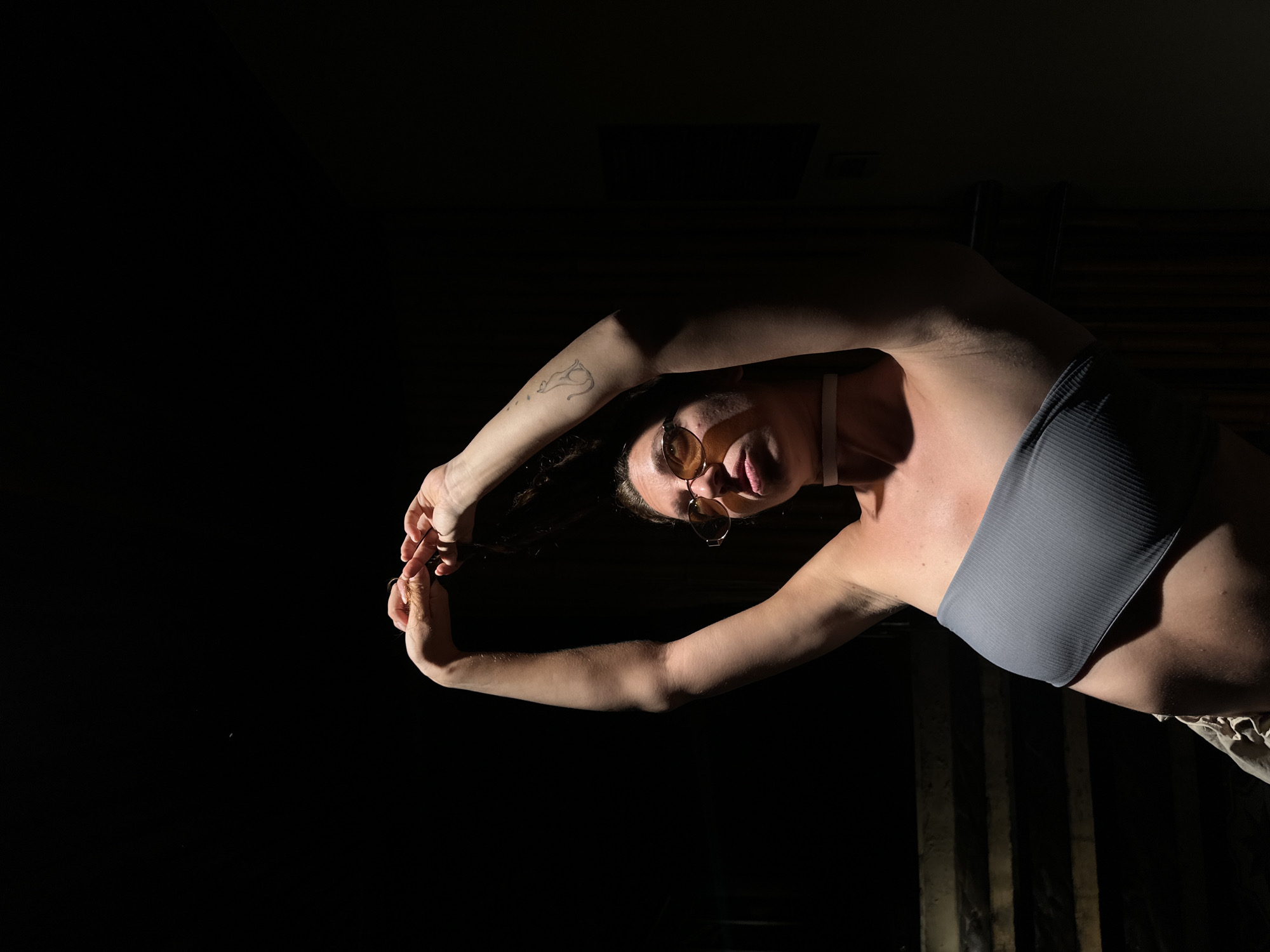 An adult female is depicted from the mid-torso upwards, positioned against a predominantly dark, unlit background. She is illuminated by a stark, focused light source originating from the upper right, casting strong shadows on her left side. Her arms are raised above her head, forming an arch, with her hands clasped or intertwined, interacting with her dark, pulled-back hair.
The subject wears yellow-tinted, oval-framed sunglasses. Her face, primarily her right side, is brightly lit, revealing skin texture and minor blemishes on her cheek. Her lips are slightly parted. A thin, light-colored band or choker is visible around her neck. She is wearing a grey, ribbed, strapless bandeau-style top. A small, stylized tattoo resembling a cat is present on the inner upper arm of her left arm (viewer's right). Her lower abdomen and a portion of a light-colored, possibly khaki, garment are visible in the bottom left corner.
The background is almost entirely black, with subtle, shadowed horizontal lines suggesting steps or a dark architectural feature visible in the lower left quadrant. The strong contrast between the intensely lit subject and the deep shadows of the environment creates a dramatic effect. No discernible elements in the image suggest a specific outdoor city location like Mù Cang Chải, Vietnam; the setting appears to be an indoor space with controlled, directional lighting.