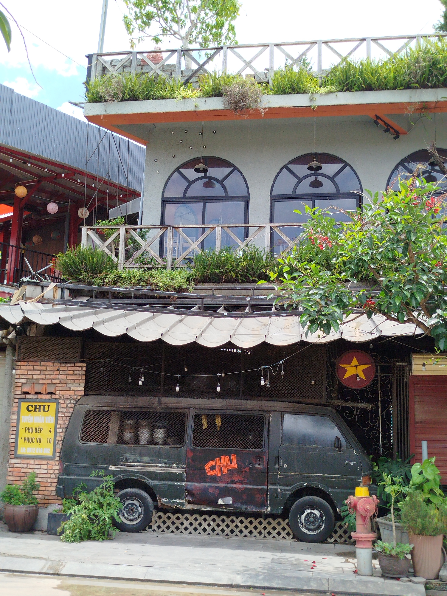 This daytime photograph captures the exterior of a multi-story building, likely a cafe or restaurant named "CHU," in Da Lat, Vietnam. The establishment features an eclectic, rustic design with a light grey facade accented by orange trim and wooden railings. Lush green plants adorn the balconies on both the mid and upper levels, creating a vibrant, natural aesthetic.
A weathered, dark green van, exhibiting rust and bearing the orange spray-painted text "CHU" on its side, is parked beneath the mid-level balcony, partially sheltered by a scalloped awning. Its windows are covered with a mesh. Adjacent to the van, a yellow sign on a brick pillar displays Vietnamese text: "CHU TUYỂN NHÂN VIÊN" (CHU Recruiting Staff), with job listings for "* PHỤ BẾP 4" (Kitchen Helper 4) and "* PHỤC VỤ 10" (Server 10), along with a contact number "LK 0912 816 892". A circular red sign featuring a prominent yellow star, reminiscent of the Vietnamese flag, hangs nearby.
String lights with bare bulbs are strung under the awning and around the van, suggesting evening use. A small tree with red blossoms partially obscures the right side of the building. The sidewalk features a red fire hydrant with a yellow top and various potted plants. No people are visible, rendering the scene quiet and still under clear, bright daylight. The adjacent building on the left has a red corrugated metal roof.