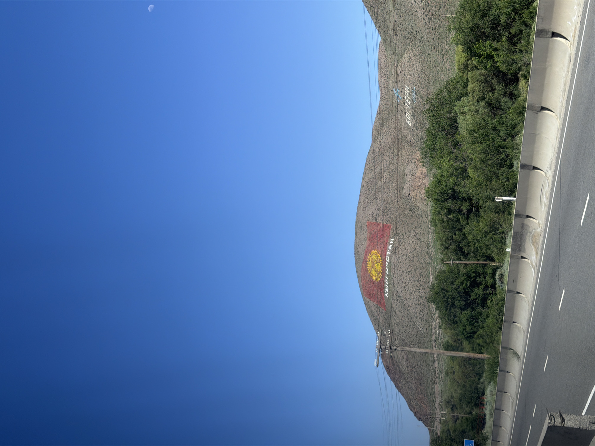 The image captures a mountainous landscape in Kyrgyzstan, as indicated by the visible text "КЫРГЫЗСТАН" and a large rendition of the Kyrgyz flag painted on the mountainside. The flag, featuring a yellow sun with a stylized crown in the center, is prominently displayed on the textured, arid slopes of the mountain. To the right of the mountain, a paved road runs alongside, bordered by a concrete barrier and lined with vegetation. The scene is bathed in bright sunlight, suggesting a clear day, likely mid-morning or afternoon given the strong shadows and the high position of the moon, which is faintly visible in the bright blue sky. Power lines and poles are scattered across the landscape, indicating infrastructure development in the area. The overall impression is one of a grand, natural setting with human elements like the road, flag, and text art integrated into it, showcasing national pride or a landmark. The lack of people suggests a focus on the landscape and its features.