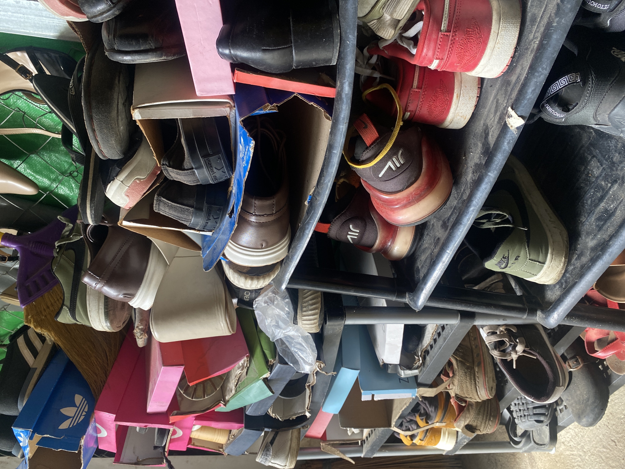 The image presents a densely packed and cluttered multi-tiered shoe rack filled with various types of footwear and storage items. The perspective is from slightly above, looking down into the shelves.
The main subjects are numerous shoes of different styles, colors, and conditions, along with metal shelving units and various cardboard boxes. The footwear includes sneakers, sandals, flats, and possibly heels, indicating a diverse collection. Many shoes appear well-worn.
No specific activity is taking place; the scene depicts stored items. The setting appears to be an indoor storage area, possibly a closet, garage, or utility room, characterized by its somewhat disorganized nature.
Notable details include:
- A blue Adidas shoe box is visible towards the bottom left, featuring the iconic Adidas trefoil logo.
- Red Jordan sneakers, identifiable by their prominent Jumpman logo, are seen towards the top right.
- A green Nike sneaker with a black swoosh is positioned on an upper shelf.
- Black sneakers with "P.U.A. FASHION" written on their heels are visible on the left side.
- A dark sandal or shoe has text that appears to be "TOKOUM" or "TOKOUMPH" on its strap.
- Brown and red sneakers display a white mark that resembles "JIL" or "J1L".
- A light blue box shows faded text that might be "NZ".
- Several torn cardboard boxes are used for storage or are simply discarded within the rack.
- A brown, fibrous broom head is wedged among shoes on the left.
- Plastic bags are also visible, adding to the cluttered appearance.
The shoes are crammed tightly onto and between the shelves, suggesting limited storage space. The lighting is consistent with an indoor environment, but the time of day is not discernible. No people are visible, and there are no direct cues about the location, other than the provided context of Davao City, Philippines.