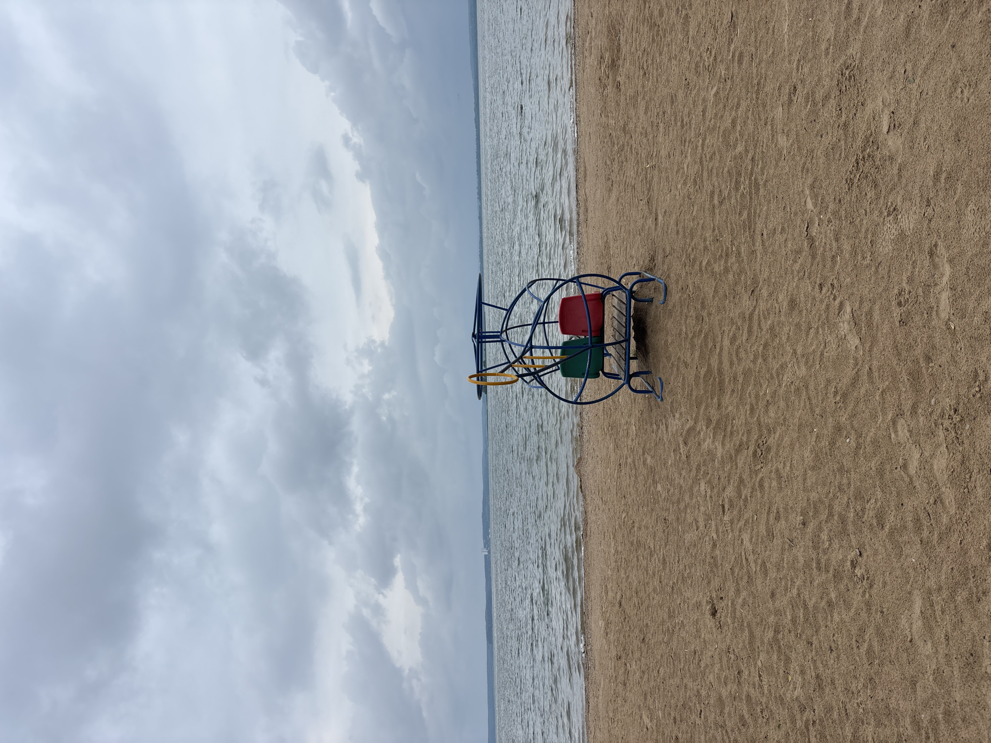 The image depicts a beach scene with a playground apparatus in the foreground. The apparatus, a blue metal structure with two seats (one red, one green) and a yellow circular frame, is positioned on the sand. The sand shows the imprints of footsteps. In the background, a large body of water, likely a lake, stretches to the horizon. The sky is overcast with heavy, grey clouds. A distant shoreline with trees and a few indistinct structures is visible across the water. The location is identified as the beach area in the village of Zhdanovichi, within the Zhdanovichi rural council, Belarus.