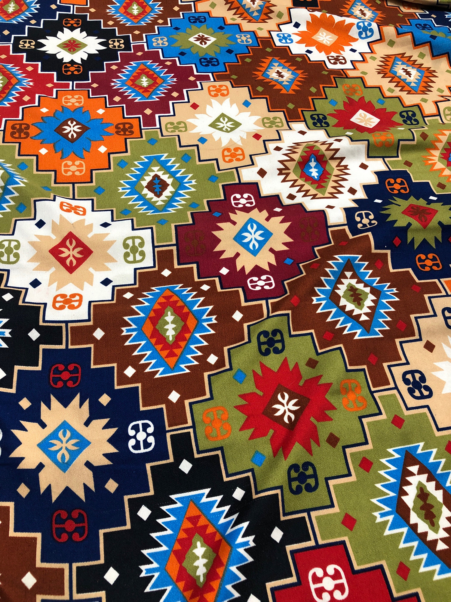 The image displays a close-up, overhead view of a patterned fabric, likely a textile or upholstery. The pattern is geometric and features a repeating motif of stylized diamond shapes and zig-zag borders. The colors are vibrant and include shades of red, orange, blue, green, brown, black, white, and tan. Small, abstract symbols resembling stylized figures or knots are also integrated into the design. The fabric appears to be slightly creased and textured, suggesting it might be a natural fiber or a blend. There is no discernible connection to the city of Larkana, Pakistan, in the visual content of the image.
