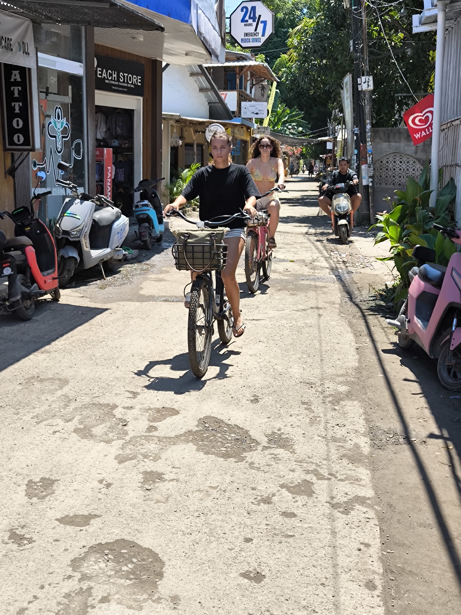 The image captures a street scene in what appears to be a tourist area in Unknown, Indonesia. The foreground shows a woman in a black t-shirt riding a bicycle toward the camera. The bicycle has a basket. A woman wearing a bikini and shorts is following behind her, also on a bicycle. Behind them, a man rides a motor scooter. Parked motor scooters line both sides of the narrow, slightly rundown street. Shops and businesses are visible, including a tattoo parlor, a retail store labeled "EACH STORE," and a medical service with a "24/7" sign. Other signs such as "COFFEE STOP" and "WALL'S" are visible further down the street, suggesting a commercial environment. The street surface is uneven and has patches of dark spots. Shadows indicate a sunny day. Trees and foliage appear above and around the buildings, adding to the tropical ambiance.