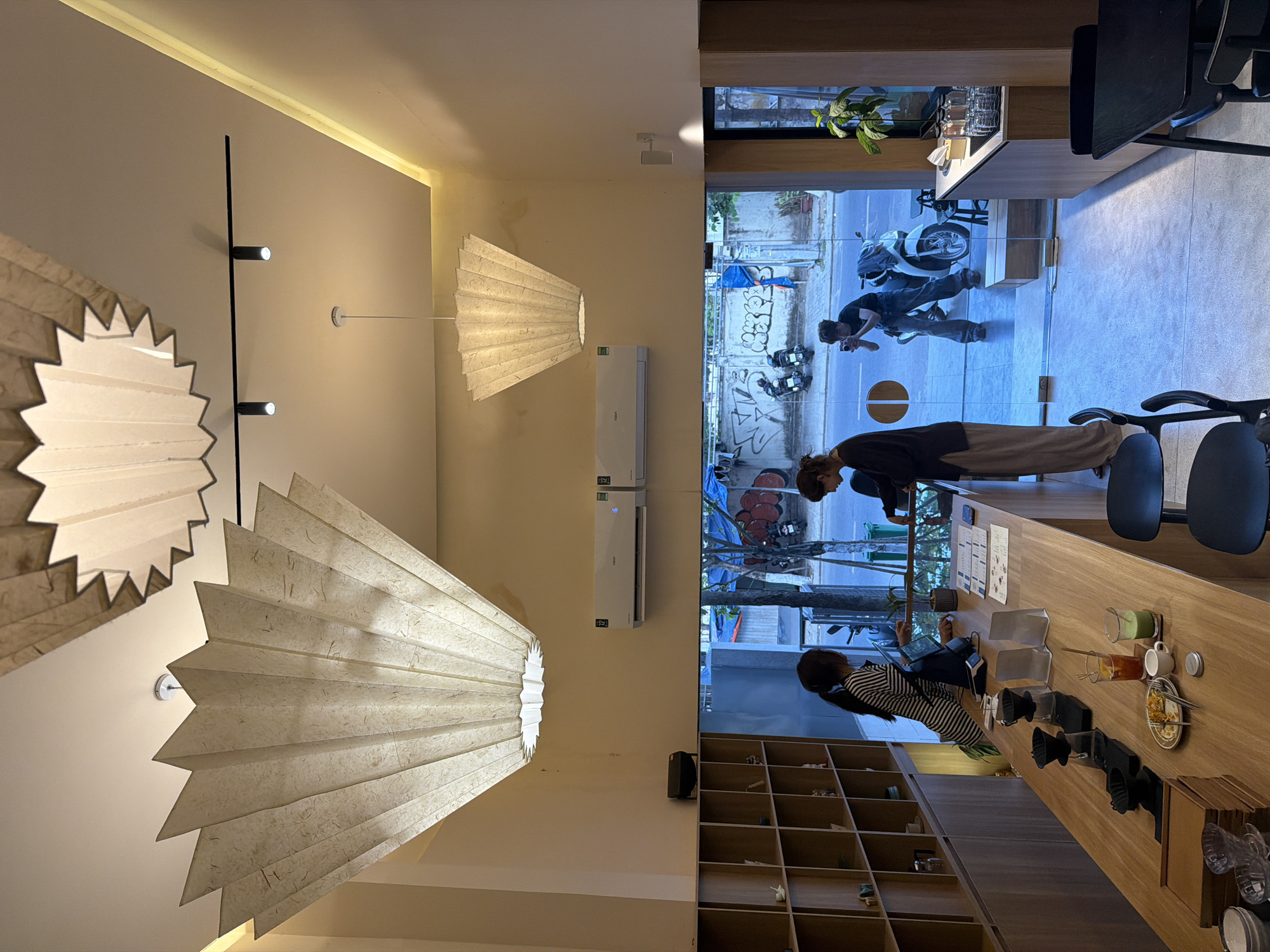 The image captures an interior view of a café in what appears to be Đà Nẵng, Vietnam, as suggested by the Vietnamese script and urban scenery visible through the large glass window.
**Environment**: The café has a modern, minimalist design. The interior is illuminated by ornate paper lanterns, one of which is large and dominates the scene. A strip of lights runs along the upper wall, and recessed spotlights provide additional lighting. A wooden shelving unit is visible, filled with various objects and plants. Outside, the scene features a typical Vietnamese street with parked motorbikes, graffiti-covered walls, and pedestrians.
**People**: Inside, there are two individuals: a woman in a striped top is sitting at a long wooden counter working on a laptop. Another person is leaning against the counter, possibly engaged in conversation. Outside, a man is taking photos of a motorbike.
**Objects**: The counter is cluttered with coffee-making equipment, cups, drinks, and a plate of food. Air conditioning units are mounted on the wall above the counter. A reflective glass window provides a view to the outside.
**Actions and Interactions**: The woman is working on her laptop, while the second person is standing and seems to be engaged in conversation or observation. Outside, the man is taking photos. The café seems to be a place of activity and relaxation.