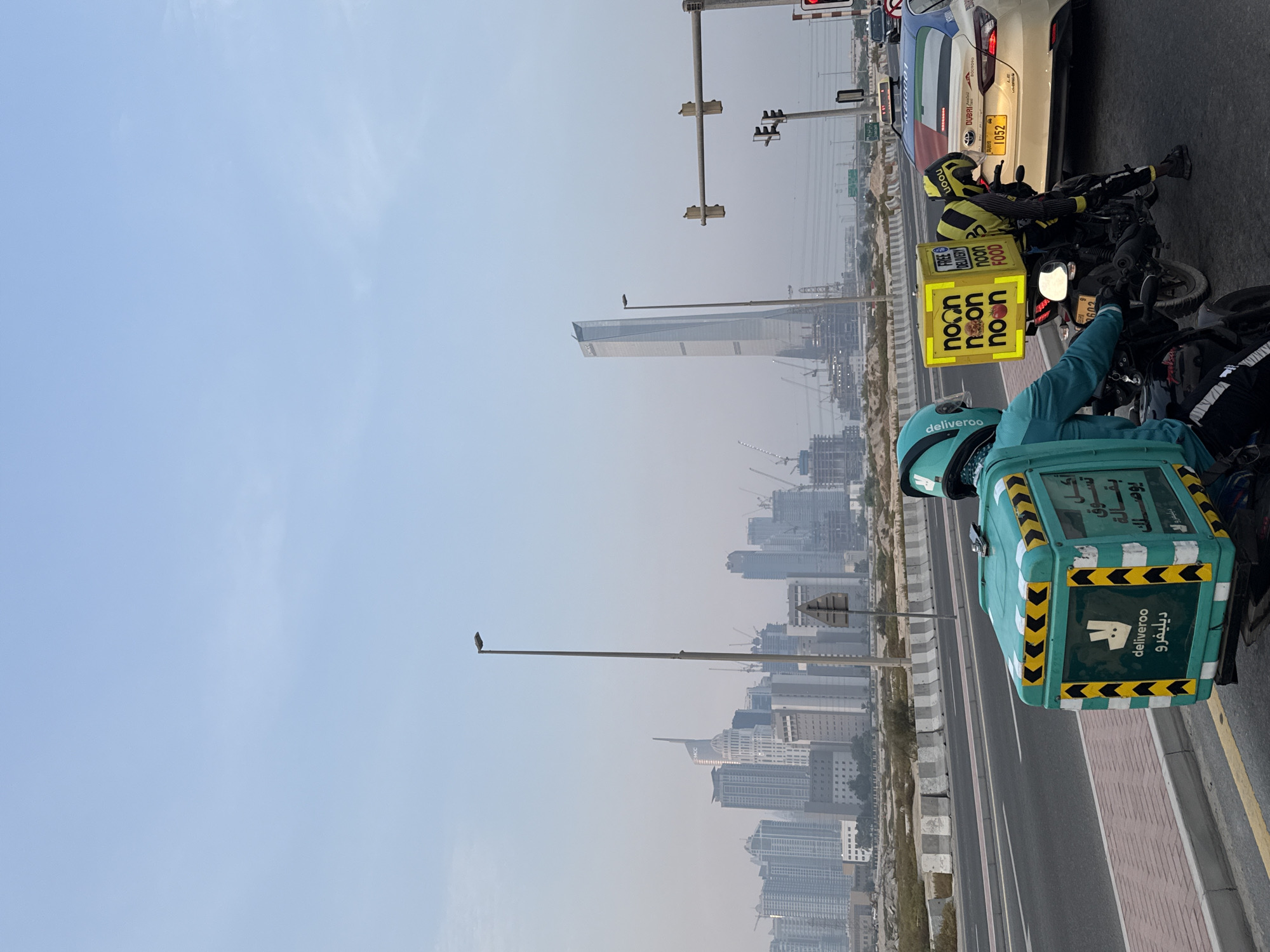 In Dubai, United Arab Emirates, a city skyline is visible in the hazy distance with numerous skyscrapers and construction cranes. In the foreground, two delivery drivers on motorcycles are stopped at a traffic light. One driver, wearing a teal helmet and jacket, is on a black motorcycle with a green and yellow Deliveroo delivery box. The second driver, wearing a yellow and black outfit and helmet, is on a black motorcycle with a bright yellow "noon food" delivery box. A white taxi with the Dubai flag design is stopped behind them, and other vehicles are in traffic. The sky is overcast with a light blue hue. The road surface is asphalt with yellow and white lane markings, and a red-brick sidewalk is visible.