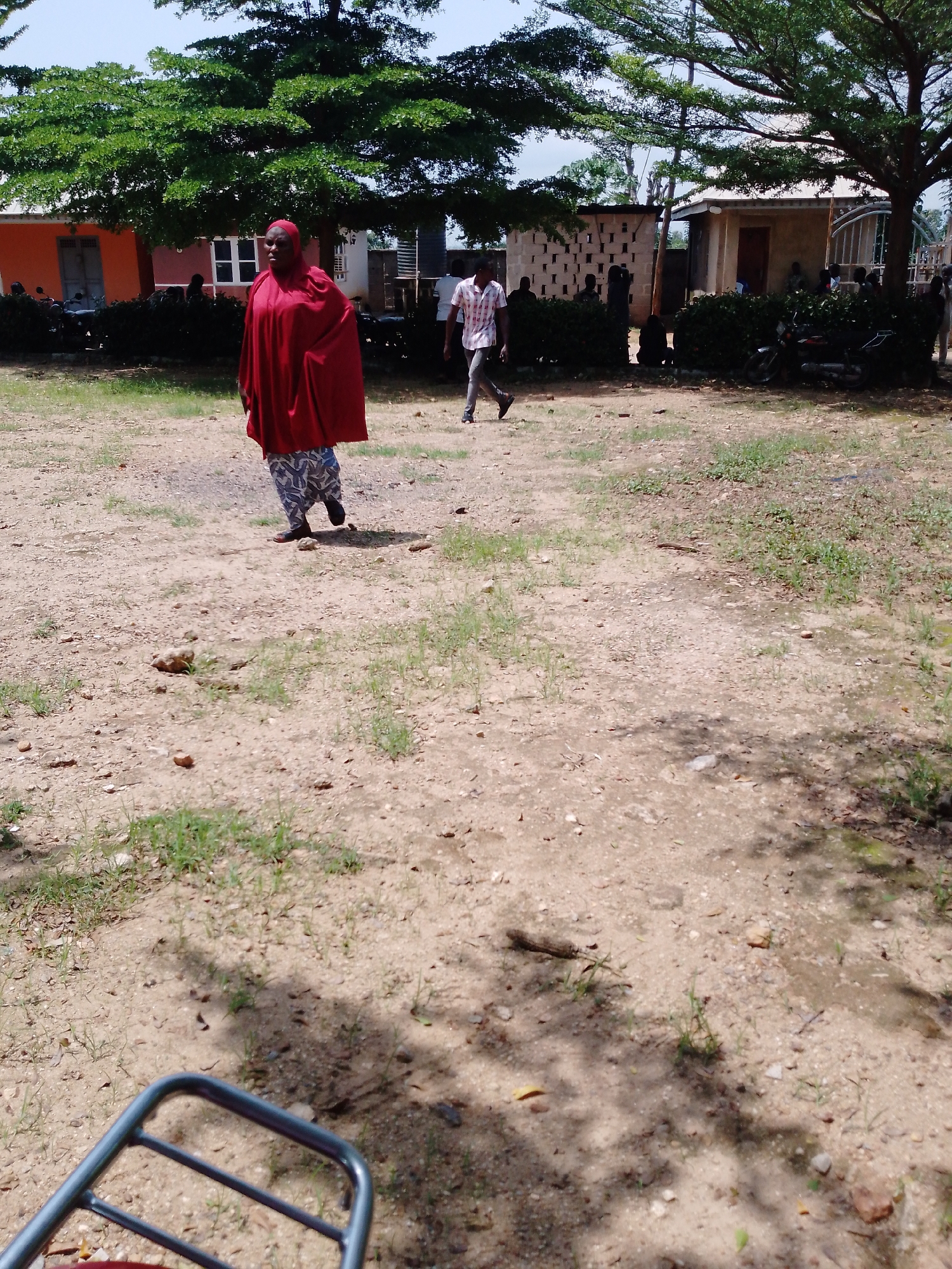 The image depicts an outdoor scene with several people and buildings under a large, leafy tree. In the foreground, a woman wearing a red hijab and a patterned skirt walks across a dusty, uneven ground. Behind her, a man in a checkered shirt and gray pants is also walking. In the background, there are a few buildings, one with an orange facade and another with a beige facade and a metal gate. Several motorcycles are parked near the buildings, and there are also some people standing or sitting in front of the structures. The ground is a mix of dirt and sparse grass. The scene appears to be set during the day, with bright sunlight casting shadows. The overall atmosphere seems casual and informal, possibly a community area or a gathering place. No text is visible in the image. The location context provided is Akwanga, Nigeria.