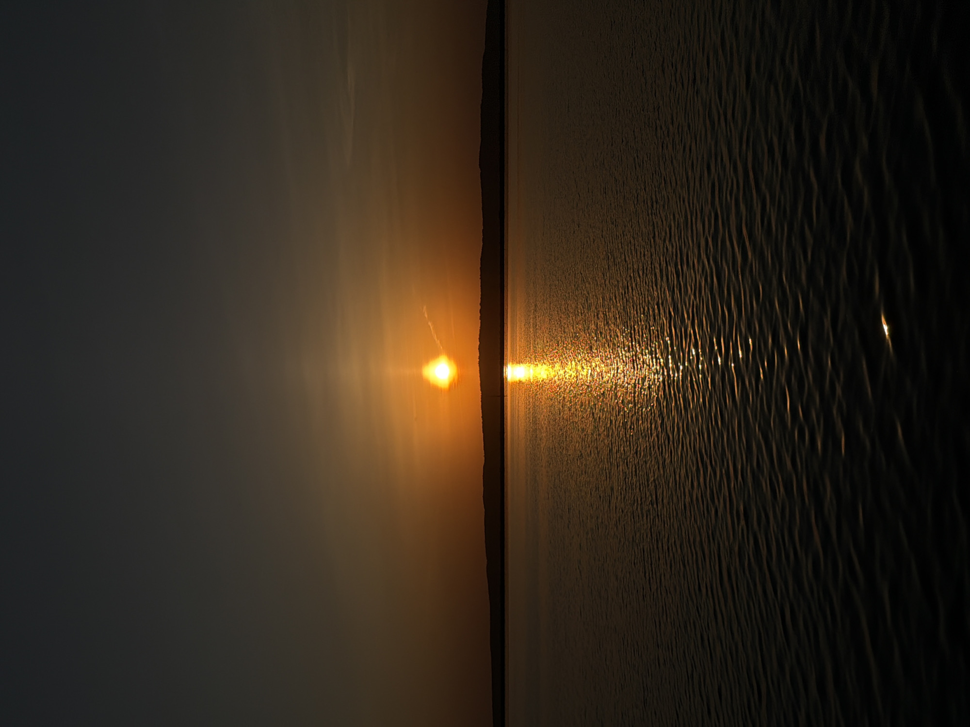 The image shows a sunset over water. The sun is a bright orange orb near the center, and its reflection stretches across the water's surface. On the left, the sky is dark. The water appears slightly rippled, creating reflections and highlights. A thin strip of land is visible between the sky and water near the sun. The photograph may be taken in the Chaikovsky region, Russia, but further details are not available in the image.