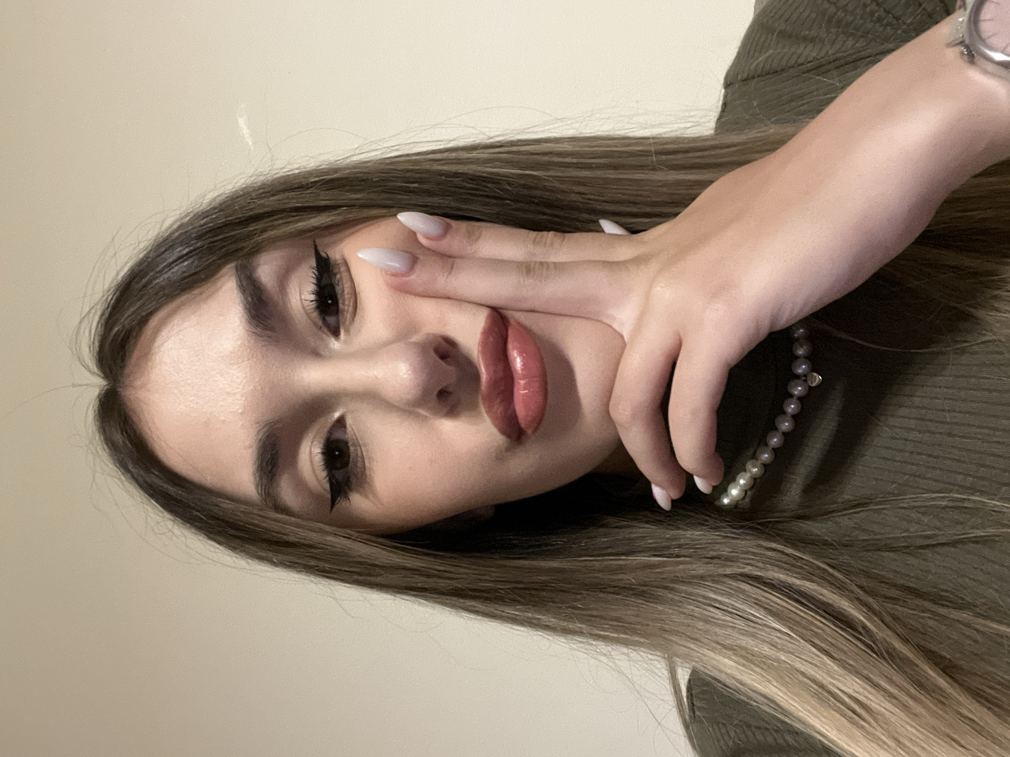 A close-up portrait, likely a selfie, features a young woman taken indoors in Sofia, Bulgaria. The image is oriented such that her head is to the right, with her body extending to the left. She looks directly into the camera with a confident gaze.
The woman has long, straight brown hair with lighter highlights, which frames her face and spreads around her. She wears a ribbed olive green top or sweater. Her left hand is raised, with fingers gently resting across her lower face and cheek, partially obscuring her mouth. Her nails are long, pointed, and painted a light, possibly off-white or pale pink, color. On her left wrist, a watch with a silver-colored band is visible, and a pearl necklace adorns her neck.
Her makeup includes a distinctive winged eyeliner, well-defined eyebrows, and glossy, plump lips with a light pink or nude lipstick shade. The background is a plain, light-colored wall, indicating an indoor setting with simple surroundings. The lighting is soft and appears to evenly illuminate her face, emphasizing her features. No visible text is present in the image.