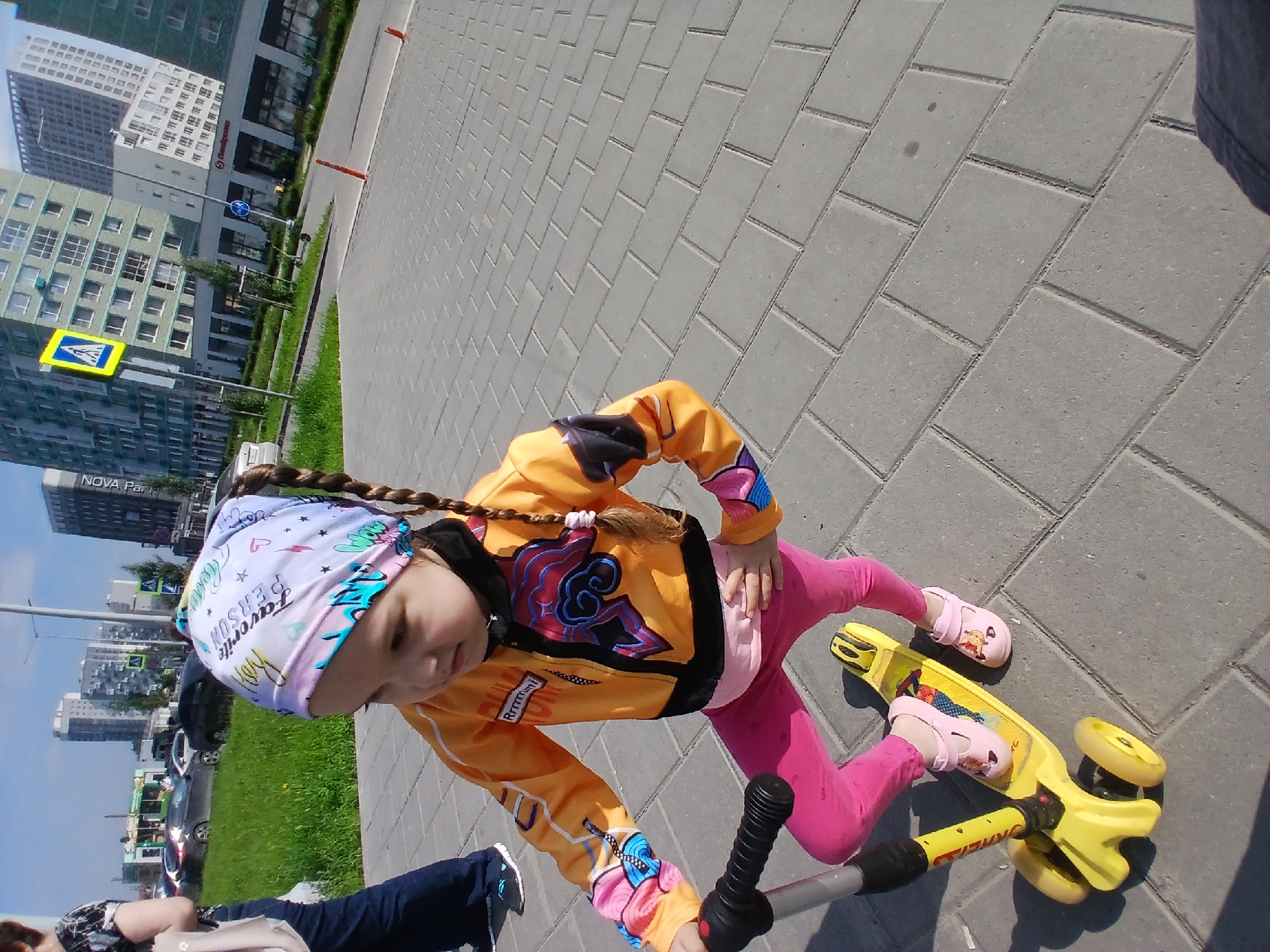 A young girl, wearing a pink bandana, an orange jacket with blue and pink graphics, and pink leggings, stands on a yellow scooter. She is outdoors on a paved walkway with gray interlocking bricks. In the background, there are modern multi-story buildings, a green grassy area, and parked cars. The sky is blue and clear. The location appears to be a residential or mixed-use area within the city of Yekaterinburg, Russia, given the architectural style and presence of a pedestrian crossing sign.