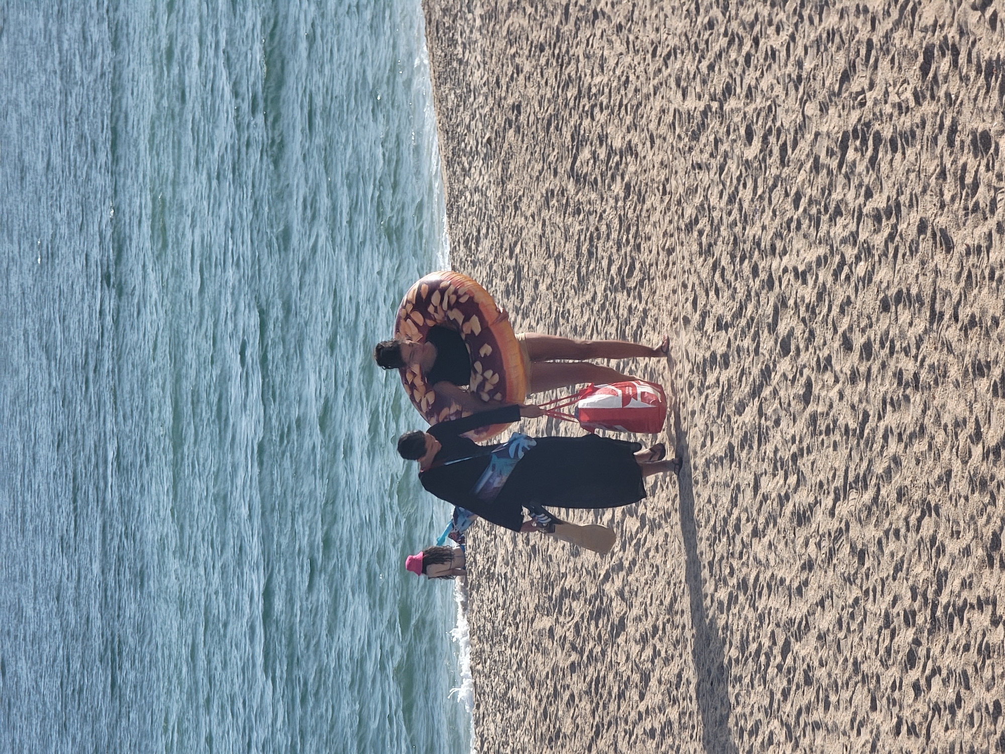 The image depicts a sunny day at a beach in Quarteira, Portugal. The scene is divided between the gentle waves of the ocean on the left and the sandy shore on the right. Two individuals are visible on the sand. In the foreground, a person wearing a dark outfit is walking, holding a long object that appears to be a surfboard or a paddle. They are walking away from the water. Behind them, closer to the water's edge, another person is lying down on a colorful inner tube, possibly a donut float. They are wearing a swimsuit and are positioned on their side, looking towards the beach. A red and white tote bag is placed on the sand near them. The sand shows textures from footprints and ripples, suggesting it is a well-used beach. The lighting indicates it is daytime, likely late morning or afternoon, with shadows cast on the sand. The water is a calm blue-green, and the sky is not visible. The overall impression is a relaxed beach atmosphere.