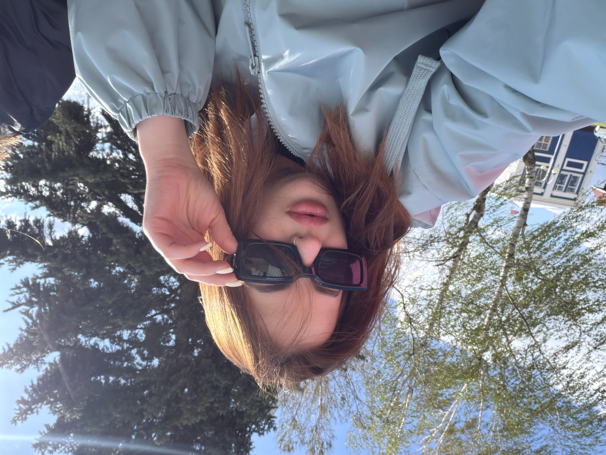 The image shows a person with long, reddish-brown hair, wearing a light blue jacket and sunglasses, in an outdoor setting. The person is holding the sunglasses slightly away from their face with their left hand. In the background, there are trees and a building with blue and white walls. The location appears to be an urban area with trees and buildings.