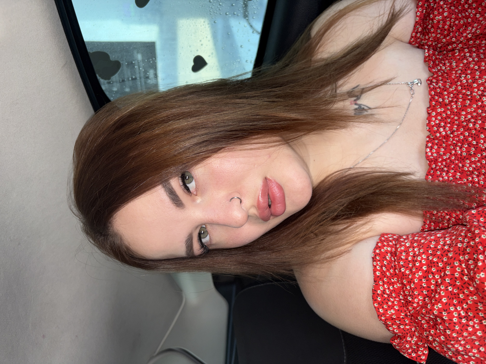 A woman is depicted seated inside a vehicle. She has shoulder-length reddish-brown hair and green eyes. A silver hoop piercing is visible in her right nostril. Her lips are full and coated with a pink gloss, slightly parted. She is wearing a red off-the-shoulder top with a small white floral pattern. A thin silver chain necklace with a small pendant rests on her chest. A dark, faded tattoo is present on her upper left chest area, partially obscured. The interior of the vehicle shows a light grey headliner. Through the window to her right, water droplets are visible, along with several dark, heart-shaped cutouts or stickers adhered to the glass. The background outside the window is blurred and appears to be an urban or suburban setting, but specific details of the city of Unknown, Unknown are not discernible. The woman is looking directly at the camera.