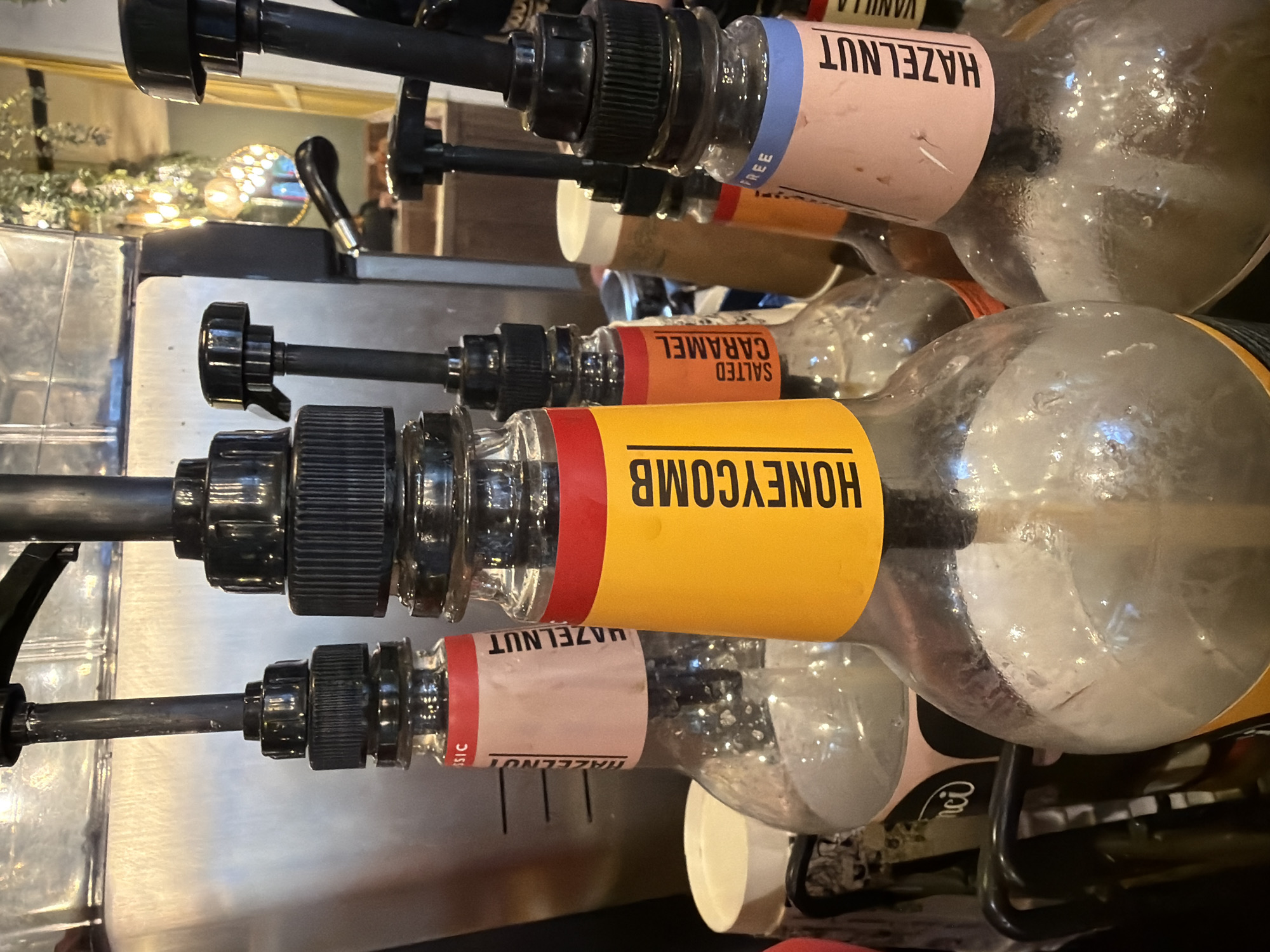 The image depicts a syrup dispensing station, likely within a cafe or coffee shop in Mole Valley, United Kingdom. Visible are four glass syrup bottles, each equipped with a dispensing pump. The syrup flavors are labeled as HAZELNUT, SALTED CARAMEL, and HONEYCOMB. The bottle labeled HONEYCOMB is the most prominent, with its yellow label and red accent. The other bottles are less focused and partially obscured. The dispensing pumps are black, and the bottles are supported by a metal frame. A stainless steel surface is visible, likely part of the counter.