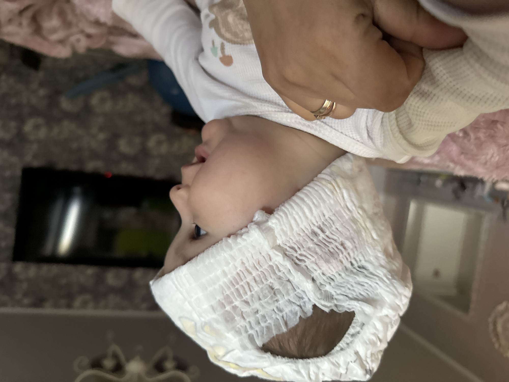 The image shows a close-up, upside-down view of a baby being dressed. An adult hand, adorned with a gold ring, is pulling a white, long-sleeved shirt over the baby's head. The baby's face is visible in profile. The background includes a patterned carpet, a pink blanket, and a television screen. The location is the interior of a home in the city of Unknown, Unknown.