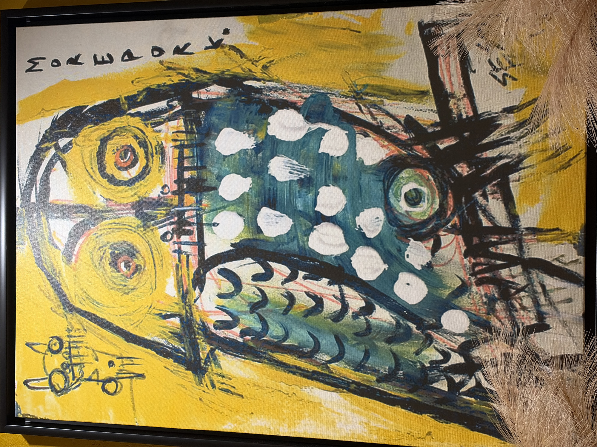 This is a close-up, medium shot of a framed, abstract painting that features a stylized fish as its primary subject. The painting is predominantly yellow, with the fish depicted in shades of teal and blue, adorned with scattered white polka dots. The fish's eye is a prominent circular element with concentric rings of green, yellow, and red.
The background is a vibrant yellow canvas with loose, expressive brushstrokes, suggesting a sense of movement or energy. Dark, jagged lines and shapes are overlaid on the fish and background, creating a sense of depth and texture. To the left of the fish, there are two large, concentric yellow circles, possibly representing additional eyes or decorative elements. Below these circles, there is a series of small, abstract black marks that appear to be a signature or inscription.
In the upper left corner of the painting, there is visible text in what appears to be a stylized font. The letters are black and run horizontally, with some characters resembling Greek letters or a phonetic alphabet.
To the right of the painting, a wispy, light-colored plume, possibly from a dried plant or decorative item, is partially obscuring the artwork. The overall mood of the painting is energetic and whimsical, with a playful yet somewhat raw artistic style. The time of day, weather, and specific location are not discernible from the image.
