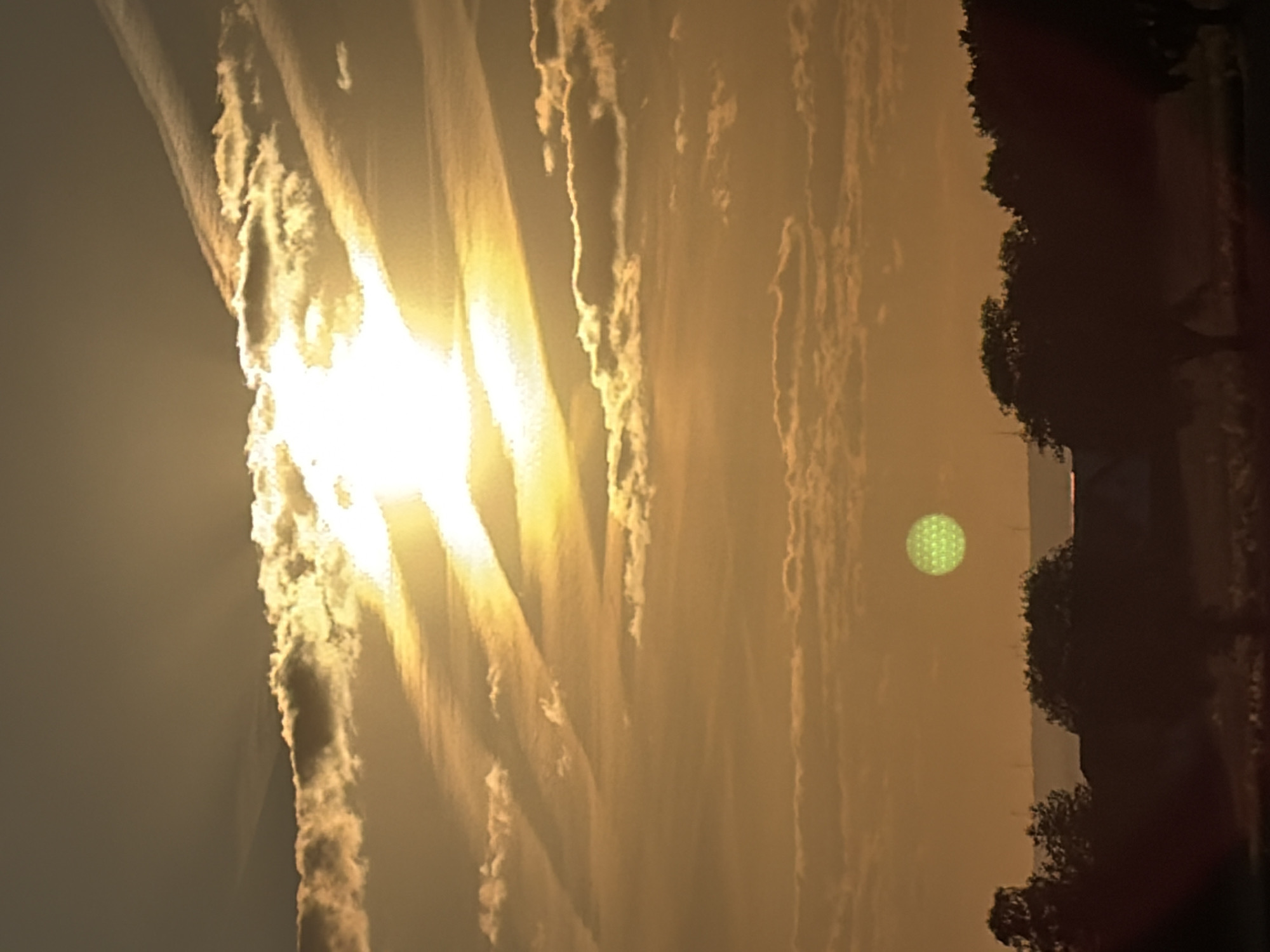 An outdoor vertical shot captures a sunset with hazy, golden light filtering through scattered clouds. A bright sunbeam shines from the top right corner, angled down towards the center of the frame, illuminating the sky with warm hues. In the middle distance, a circular object with a textured, patterned surface hovers in the sky, glowing with a faint green luminescence. Silhouetted trees and buildings are visible in the foreground against the lower horizon. The overall atmosphere is serene and visually striking.