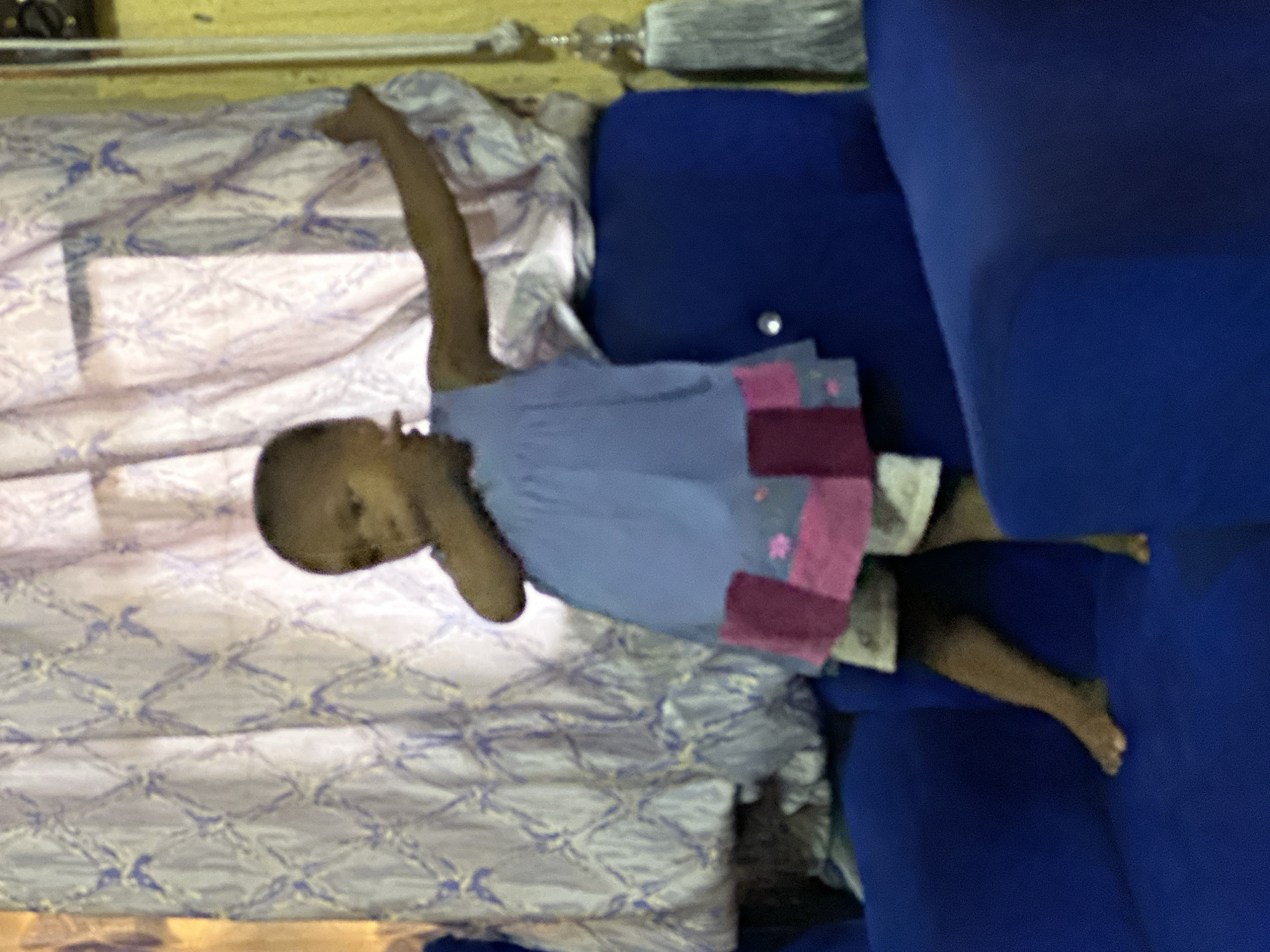 A young child with dark skin and a shaved head stands on a blue sofa, wearing a light blue dress with pink and grey fabric patches. The child's left arm is extended outwards, and their right hand is near their mouth. The child is facing towards the left of the frame. Behind the child, a light purple curtain with a patterned design is visible, partially obscuring a window. To the right of the child, a tassel hangs from what appears to be a curtain rod. The room is dimly lit, with a yellow wall visible on the far right. The overall scene suggests an indoor domestic setting. The image was taken in Narayi, Nigeria.