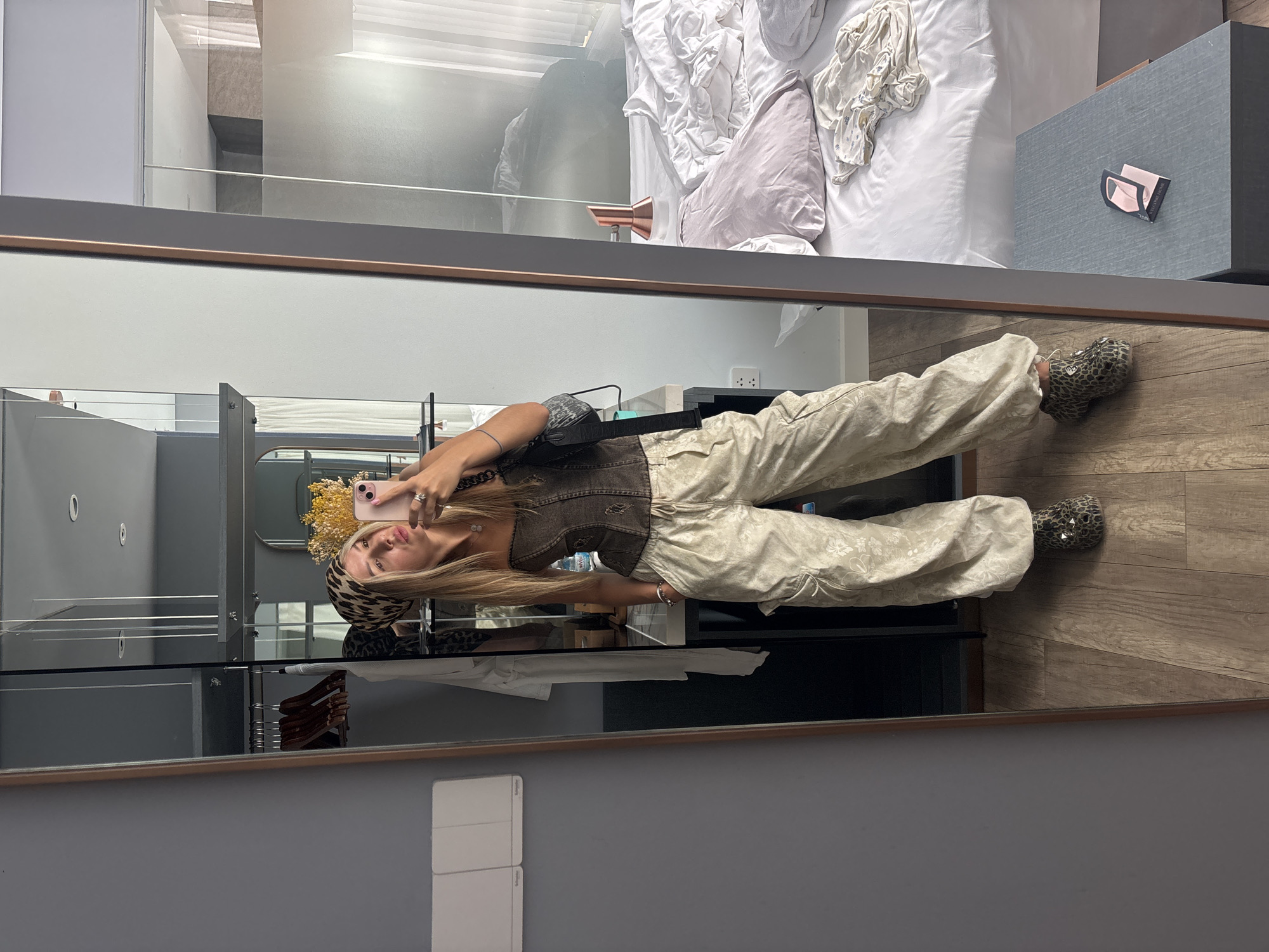 A young Caucasian woman with blonde hair is taking a mirror selfie in a hotel room. She is wearing a brown corset top, baggy tan cargo pants with a floral print, and leopard-print shoes. She has a leopard-print headscarf on and is holding a pink phone in her right hand. A black shoulder bag is slung across her body. In the mirror reflection, a messy bed with white and pink linens is visible, along with a bedside table with a "Welcome Home" card on it. The room appears to be a standard hotel room with a closet and a large mirror.