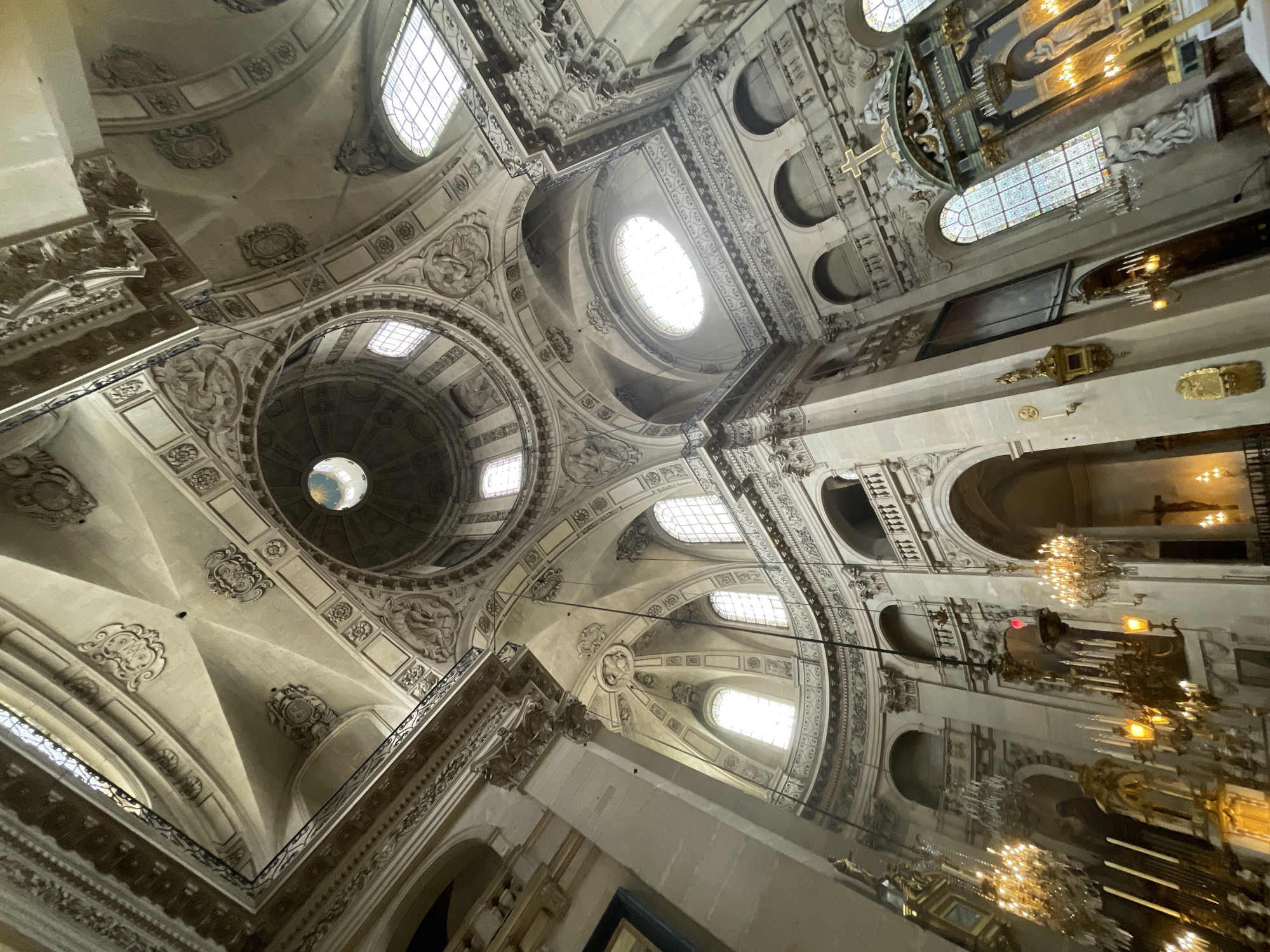 This is an interior view of the dome and upper walls of the Saint-Roch Church in Paris, France. The camera is angled upwards, capturing the grandeur of the architecture. The dome features intricate decorative moldings and is illuminated by a circular opening at its apex, through which a glimpse of a blue sky is visible. Several arched windows, some with stained glass, punctuate the walls of the nave and galleries. The interior is adorned with classical architectural elements, including columns, pilasters, and sculptures. Chandelier-style light fixtures hang from the ceiling, casting a warm glow on the ornate decorations and marble elements, including an altar with a golden cross and statues. The overall impression is one of rich historical and religious significance.