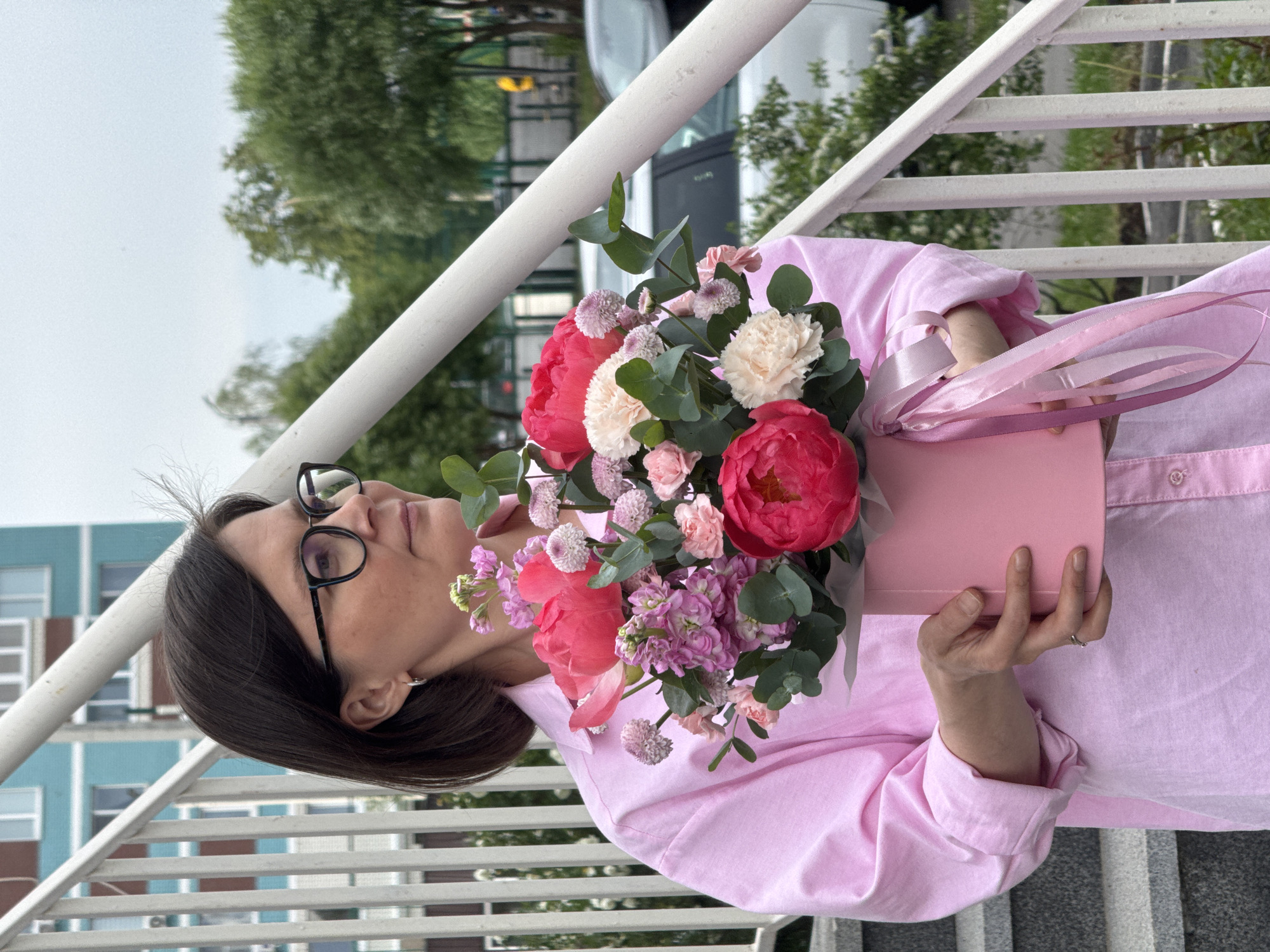 A woman with dark hair and glasses holds a pink floral arrangement in a pink box with pink ribbons while standing on a staircase. The staircase has white railings. The woman is wearing a pink button-down shirt. In the background, there are trees, buildings, and a vehicle. Based on visual cues such as the building architecture and color palette, this photograph was likely taken in the city of Izhevsk, Russia.