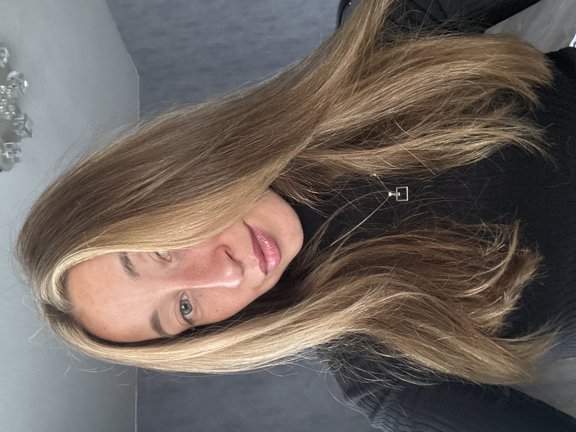 A woman with long, blonde hair and blue eyes looks directly at the camera, her head slightly tilted. She is wearing a black turtleneck sweater and a silver necklace with a small, square pendant. The background is a textured gray wall. A ceiling light fixture with crystal elements is visible in the upper right corner of the image. The image appears to be a selfie taken indoors. There is no indication of the city of Chaikovsky, Russia, in this image.