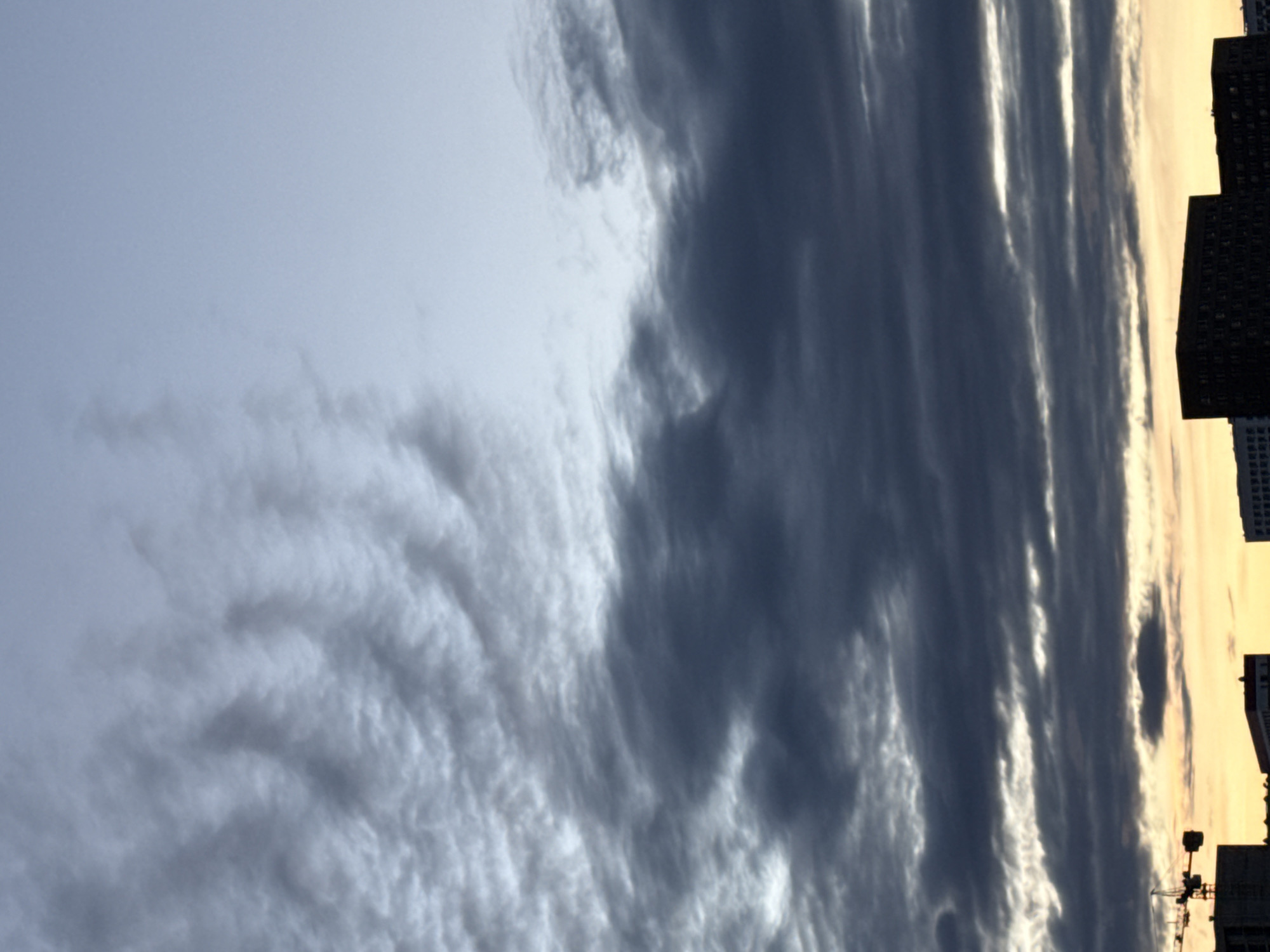 The image shows a cloudy sky in Moscow, Russia, with buildings visible along the right edge of the frame. The sky is mostly covered with various types of clouds, ranging from light, striated clouds to darker, denser formations. The lighting suggests either dawn or dusk. A construction crane is also visible among the buildings.