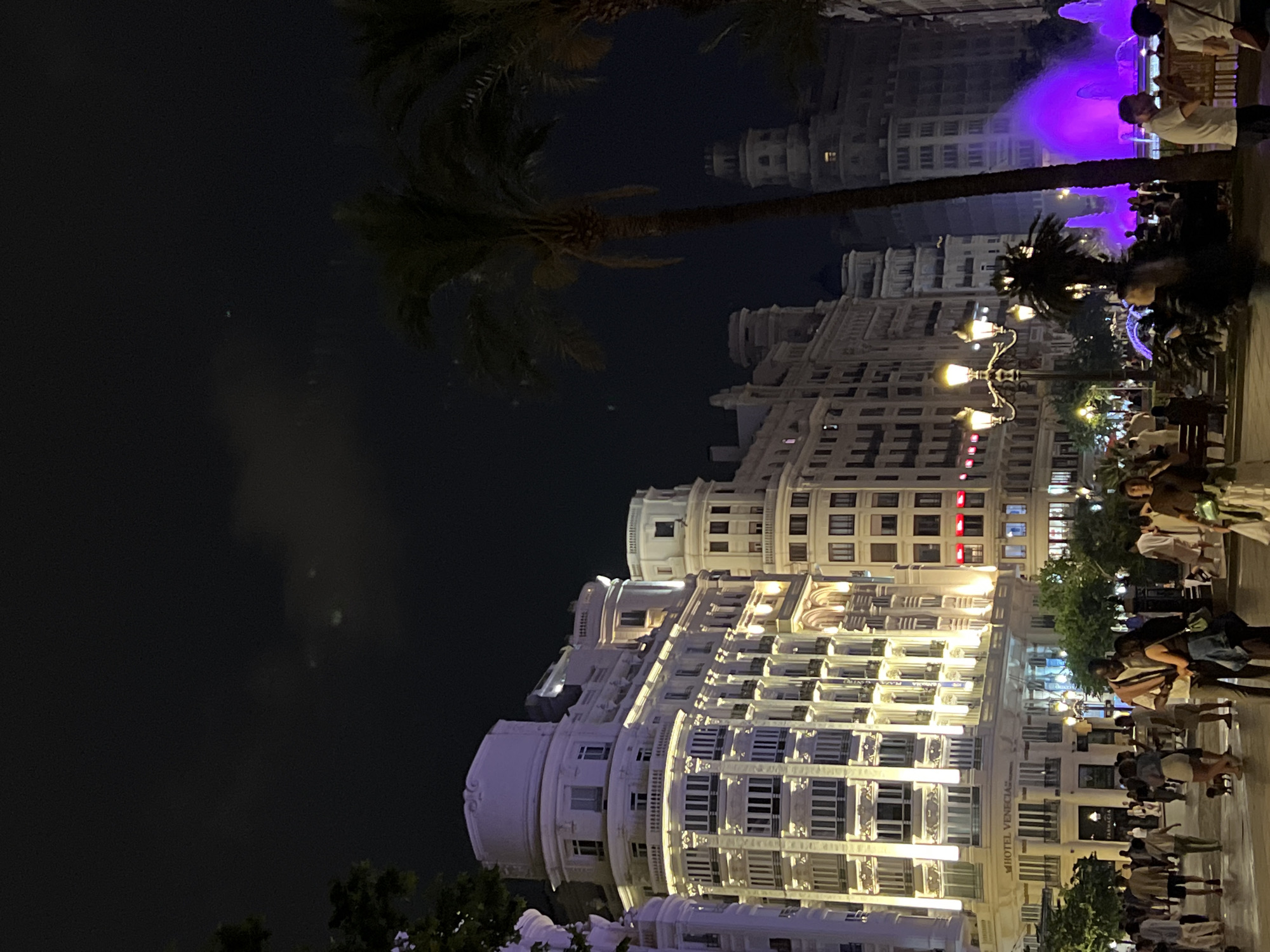 The image captures the illuminated Hotel Venecia in València, Spain at night. The hotel's white facade is brightly lit, showcasing its architectural details. Palm trees frame the building, adding a tropical element to the urban scene. Pedestrians are visible on the sidewalk in front of the hotel, some standing and others walking. Street lamps provide additional illumination. In the background, a purple-lit fountain can be seen. The dark night sky contrasts with the bright lights of the building and surrounding area.