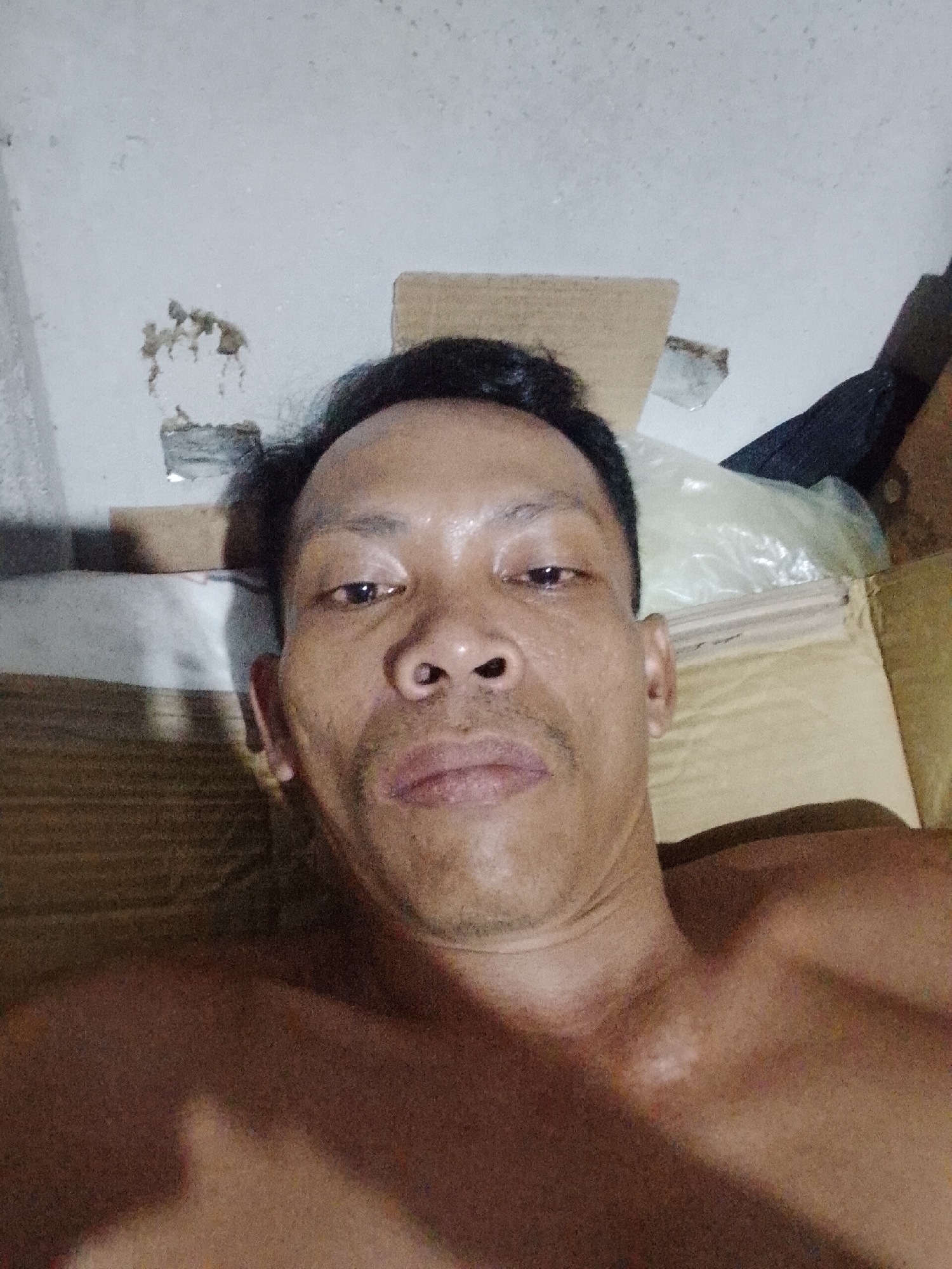 A close-up selfie of a shirtless adult male with dark hair and a mustache, lying down. The background is a wall with peeling paint and cardboard boxes. The photo was taken indoors in what appears to be Bukit Ketok, Indonesia, based on geolocation data.