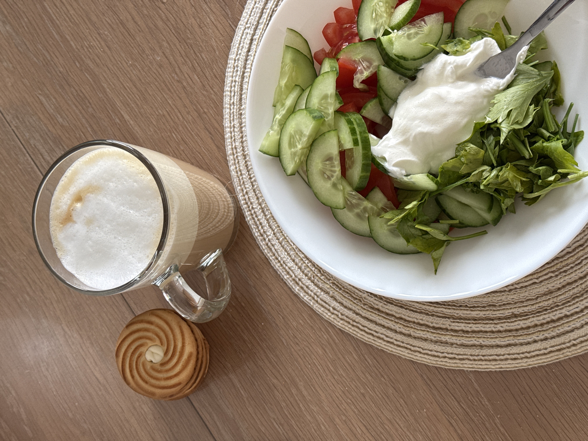 A top-down shot displays a latte in a glass mug, a round cookie, and a plate of salad with tomatoes, cucumbers, parsley, and a dollop of white cream, all arranged on a wooden surface. A beige woven placemat sits underneath the salad plate. The latte is foamy on top. The cookie is swirled in design. The salad includes a mix of sliced tomatoes, cucumbers, fresh parsley, and a creamy white sauce. The location cannot be determined to be Dikaya Gar', Russia based on the image.
