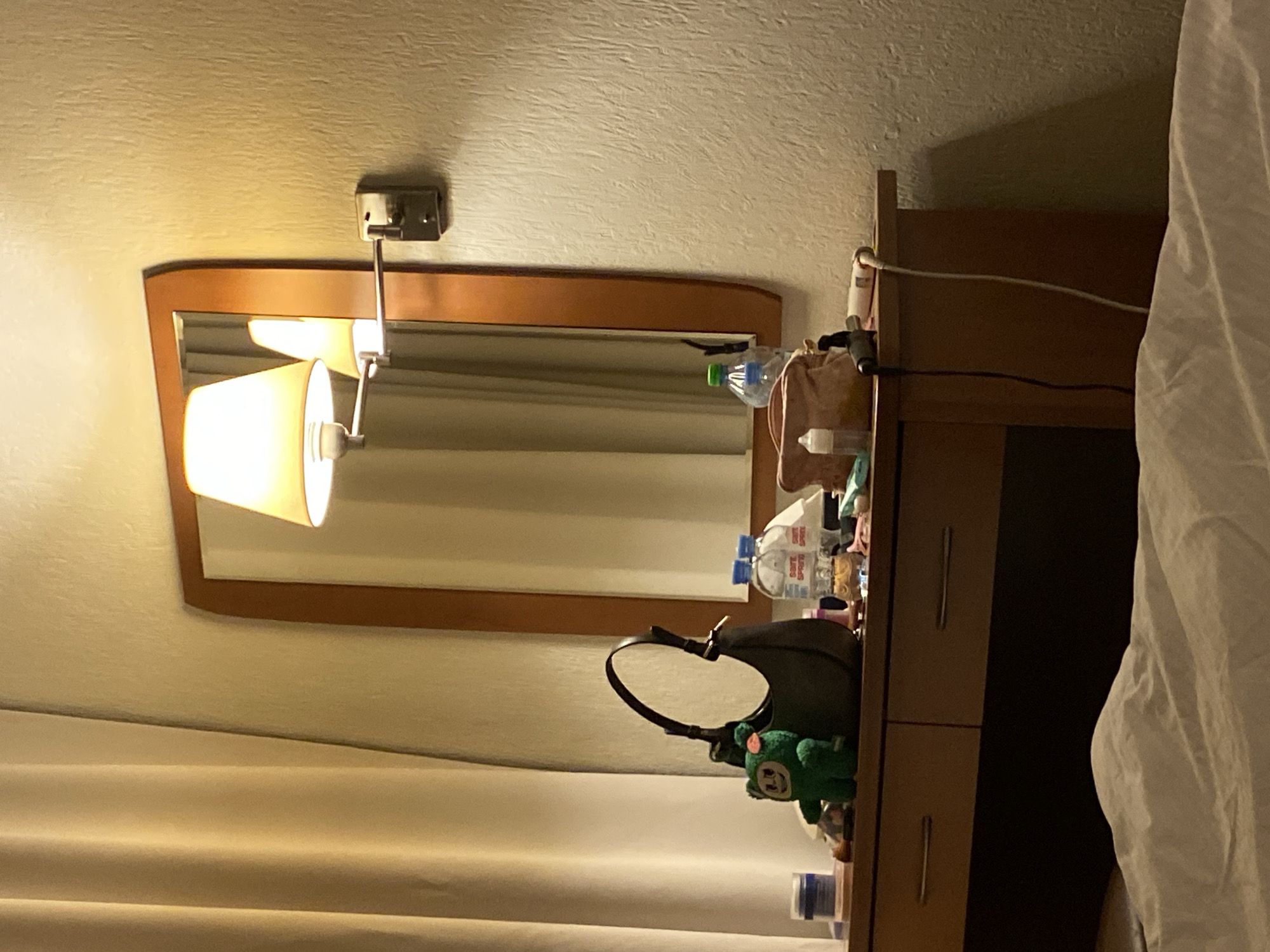 The image shows a bedside table with a mirror and a lamp in a room. On the table are several items, including a purse, water bottles, and a stuffed green animal. A curtain is visible in the background. The scene appears to be indoors in a room with warm lighting. There is no verifiable information to determine if it's located in Saint Petersburg, Russia.
