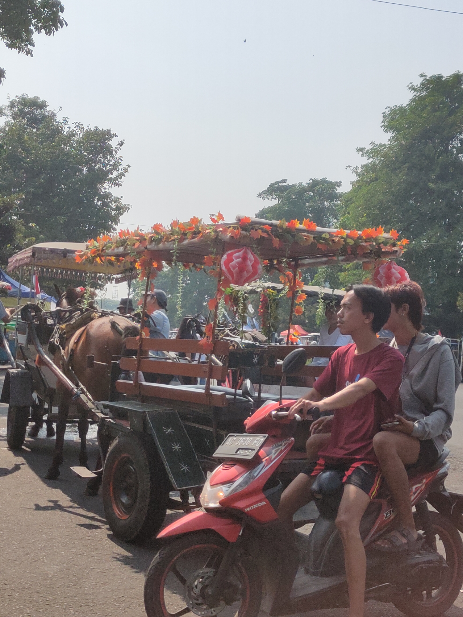 In the city of Ngasem, Indonesia, a horse-drawn carriage is pulled by a brown horse and adorned with orange and red floral decorations and paper lanterns. The carriage carries several passengers, including a man in a hat and a person wearing a blue shirt and cap. In the foreground, two individuals are seated on a red motorcycle. The person in front, wearing a red t-shirt and black and red shorts, is operating the motorcycle. The person behind, wearing a grey hoodie and black shorts, is holding a smartphone. The scene is set on an asphalt road with trees visible in the background. The sky is a pale, hazy blue, suggesting a bright, possibly warm day.