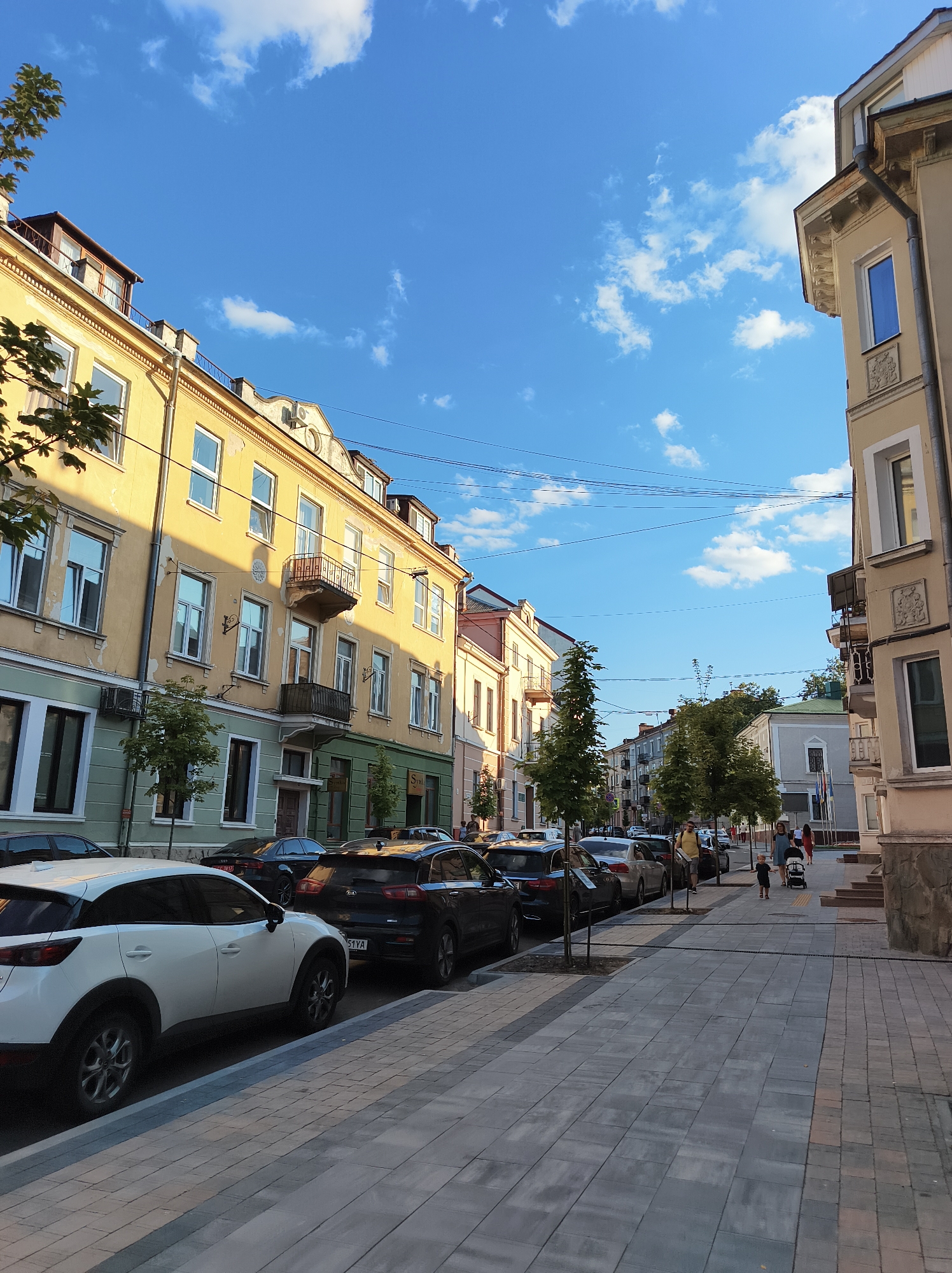 A street scene in Ternopil, Ukraine, shows buildings lining a pedestrian walkway with parked cars on the street. The sky is blue with scattered clouds. The buildings are a mix of architectural styles, with some featuring ornate balconies and yellow facades, while others are painted in lighter colors. Trees are planted along the sidewalk, providing some shade. Several cars are parked along the curb, including a white SUV in the foreground. A few people are walking on the sidewalk, including a man with a child and a woman with a stroller. The street is paved with interlocking concrete blocks, and the sidewalk is made of similar material. The overall impression is of a quiet, urban environment on a sunny day.