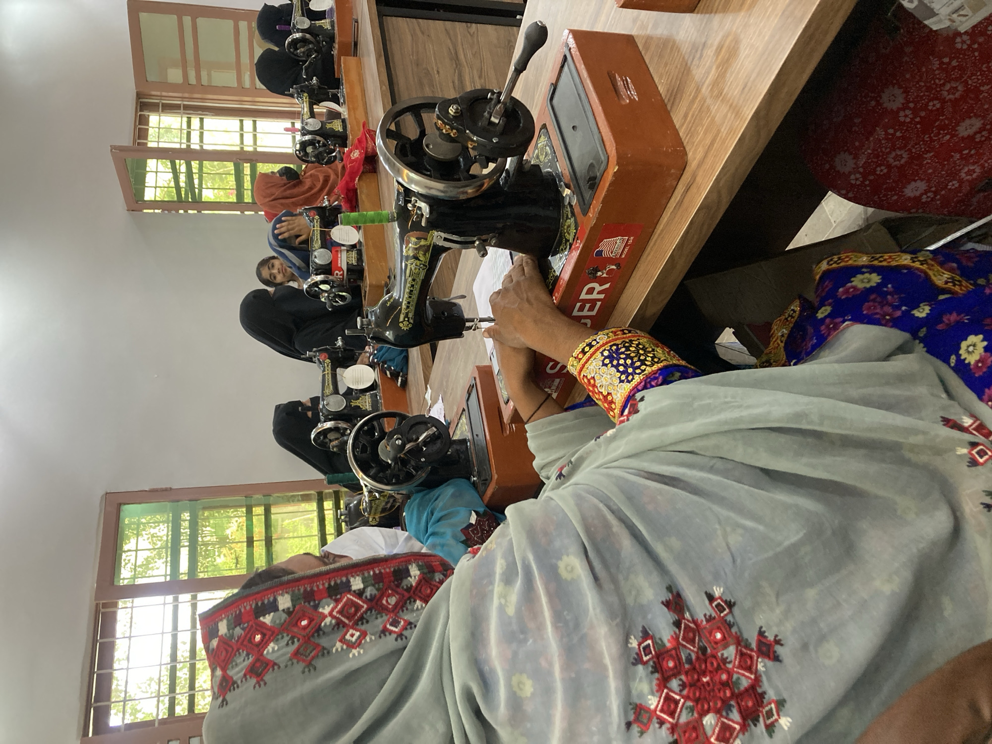 In a brightly lit room in Rāwāni, Pakistan, several women are seated at individual sewing machines along a long wooden table. The room has multiple windows providing natural light. The women, some wearing traditional head coverings, are engaged in operating the sewing machines. One woman in the foreground is dressed in a gray garment with red geometric embroidery, and a brightly colored and patterned cuff. The sewing machines are old-fashioned, likely treadle-powered models. The scene depicts a sewing workshop or class.
