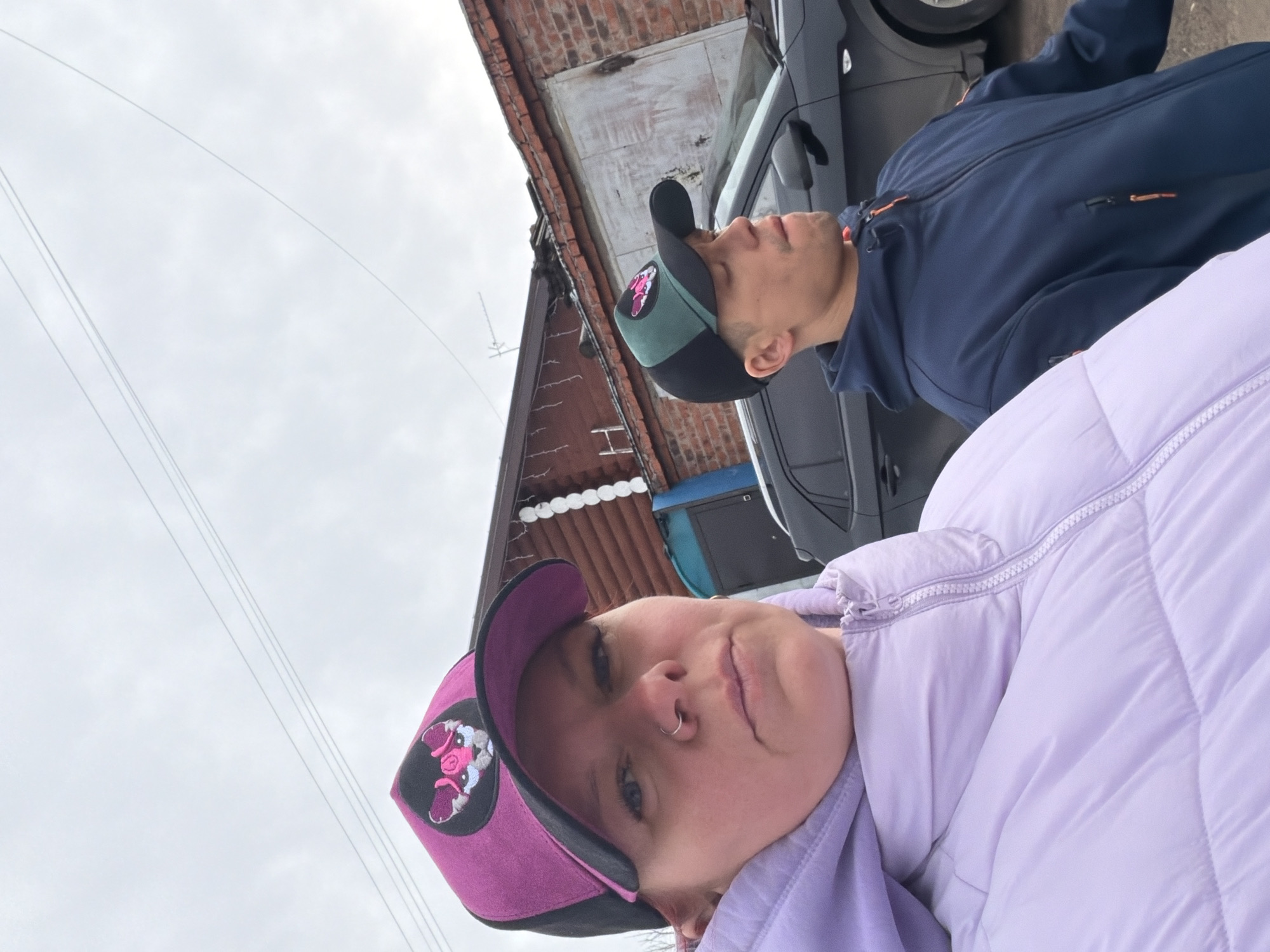 Two individuals are visible in an outdoor setting, likely in Chaikovsky, Russia. One person wears a lilac-colored quilted jacket and a purple baseball cap with a pig patch. The other person is wearing a dark blue jacket and a green and black baseball cap with a pig patch. They appear to be standing next to a gray car and in front of a brick building with a corrugated metal roof. The sky is overcast. Electrical wires are visible in the upper left.