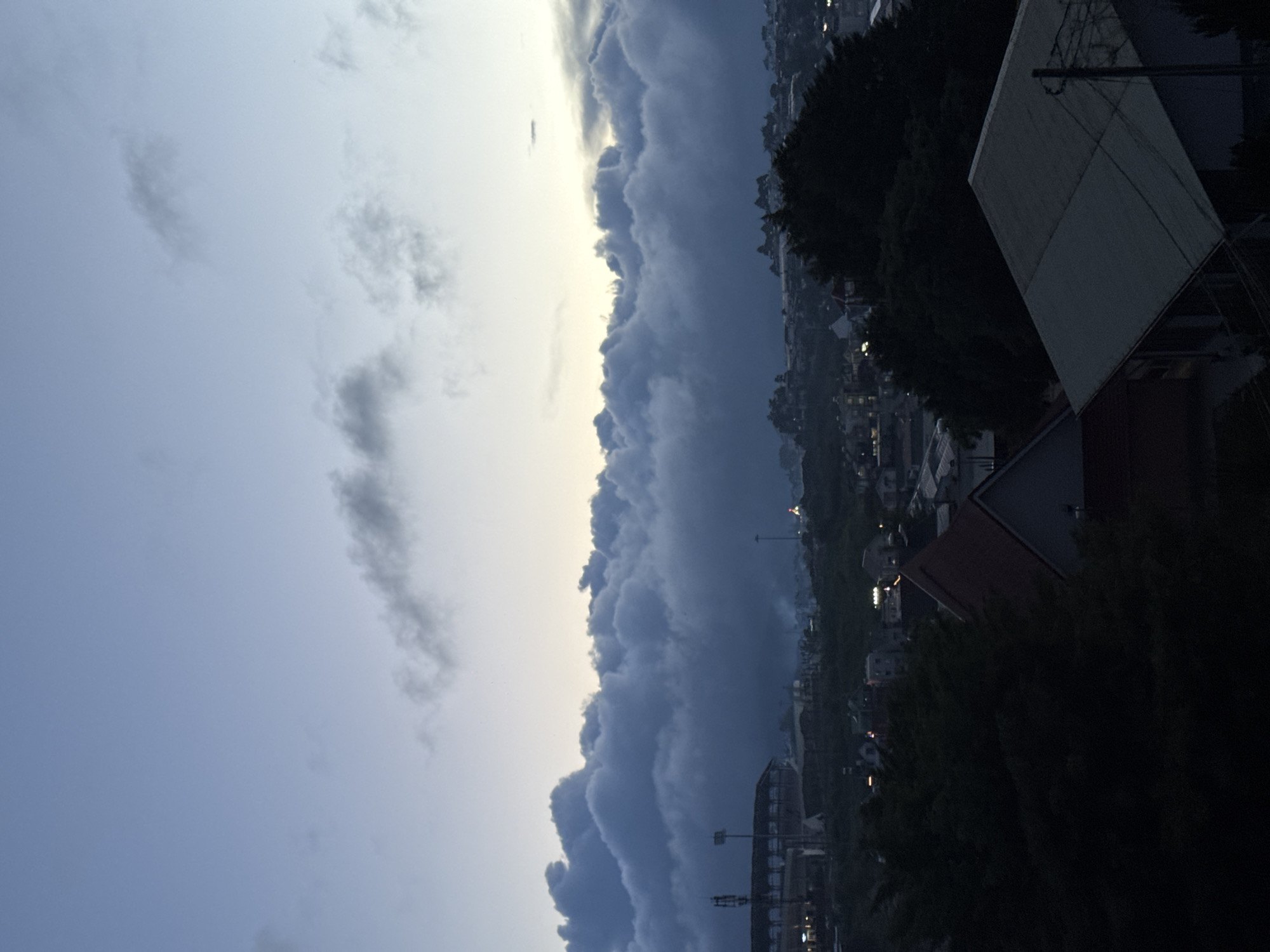 The image shows a view of a town at dusk, with the sky taking up the left side of the frame and the town on the right. The sky is mostly cloudy, with the clouds appearing dark and heavy in some areas. A break in the clouds lets some light through, creating a brighter area in the sky. On the right side of the image, there are houses and buildings visible, many of which are obscured by trees. Lights from buildings illuminate parts of the scene, indicating it's after sunset. The location is likely Lam Vien - Da Lat, Vietnam. There are no people visible in the image, only architecture and natural elements.