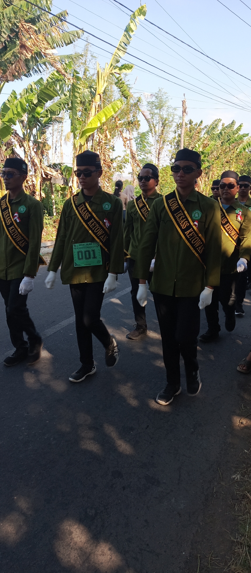 In Ketowan, Indonesia, a group of young men in uniform march in formation on an asphalt road. They wear dark green long-sleeved shirts with black pants and black skullcaps. Over their shirts, they have gold sashes with black lettering that reads "KOM. IKSASS KETOWAN". The man in the center has a green bib with the number "001" and some Indonesian text. They all wear white gloves and dark sunglasses. The sun is shining, casting shadows on the road, and the background features lush green foliage, including banana trees, and a bright blue sky. The scene appears to be a parade or formal procession.