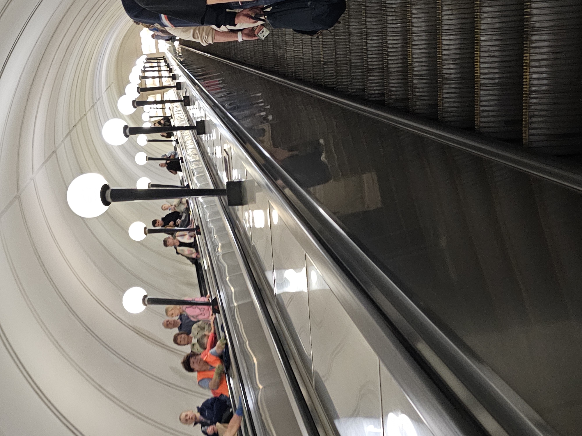 This image depicts a busy escalator in a Moscow Metro station. The escalators are wide and appear to be made of metal. Several people are seen ascending and descending the escalators. On the left escalator, a woman in an orange vest is visible, suggesting she might be a station attendant or worker. Other individuals are casually dressed, some holding bags. The station features a distinctive curved ceiling with arched lighting fixtures, a common architectural style in the Moscow Metro. The lighting is bright, with large spherical lamps casting a white glow. The perspective suggests the photograph was taken from a slightly elevated position, looking down a long, curved tunnel. The overall impression is one of a functional and well-lit public transportation hub.