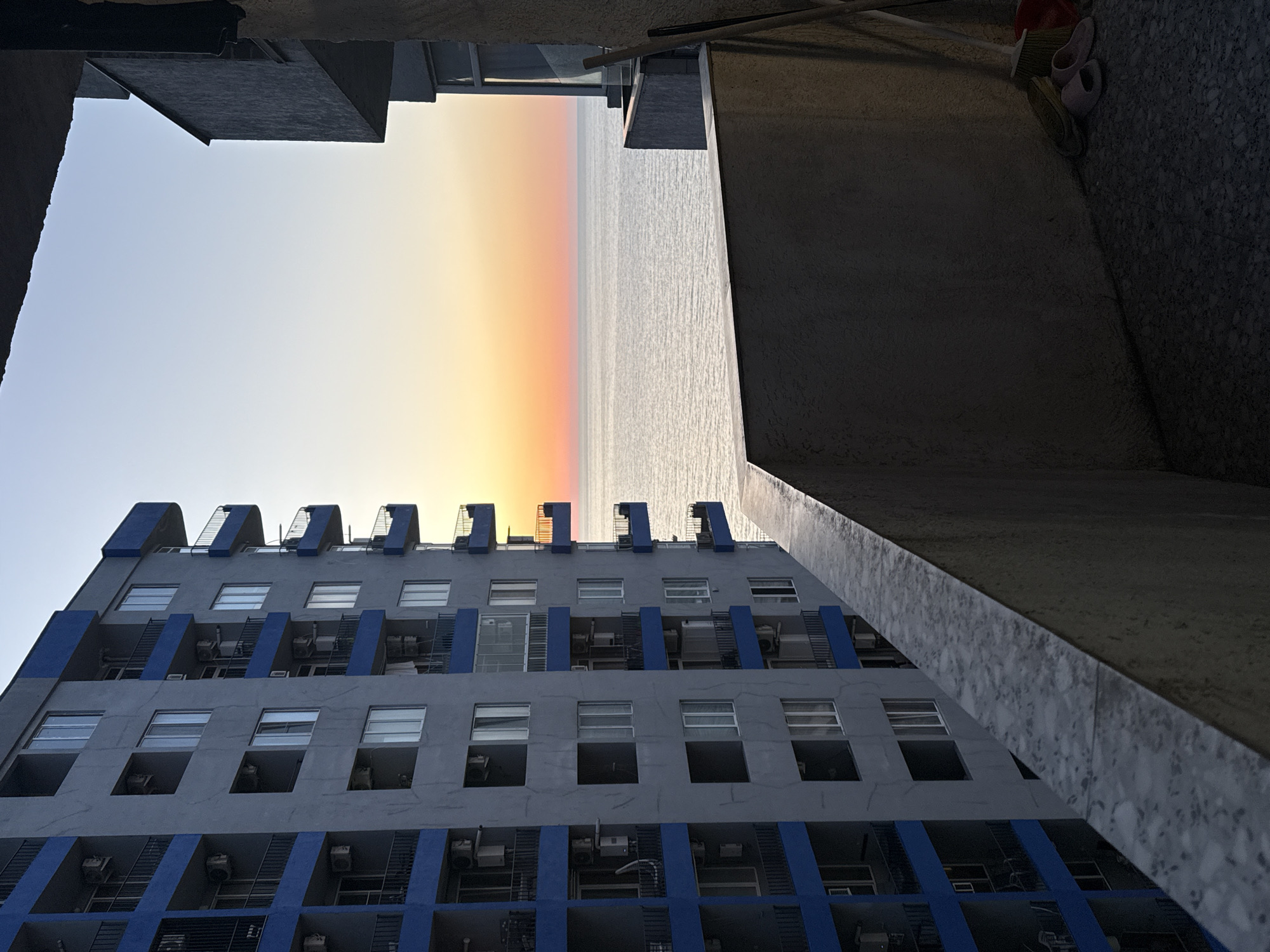 The image, taken in Batumi, Georgia, depicts a tall, gray building with blue accents, possibly an apartment complex or hotel, situated in close proximity to the Black Sea, as suggested by the horizon line dividing a bright sky and calm water. The photograph is taken from a low angle, pointing upwards between two structures, one being a concrete wall. There is a glimpse of the cityscape in the distance beyond the building. A broom and sandals are visible on the right-hand side, on the right-hand concrete wall. The lighting suggests it is either sunrise or sunset. The building has multiple stories with many windows. Small air conditioning units are located inside the windows, or beneath them on the balcony. The photograph appears to have been taken from a building's balcony or from a narrow alley.