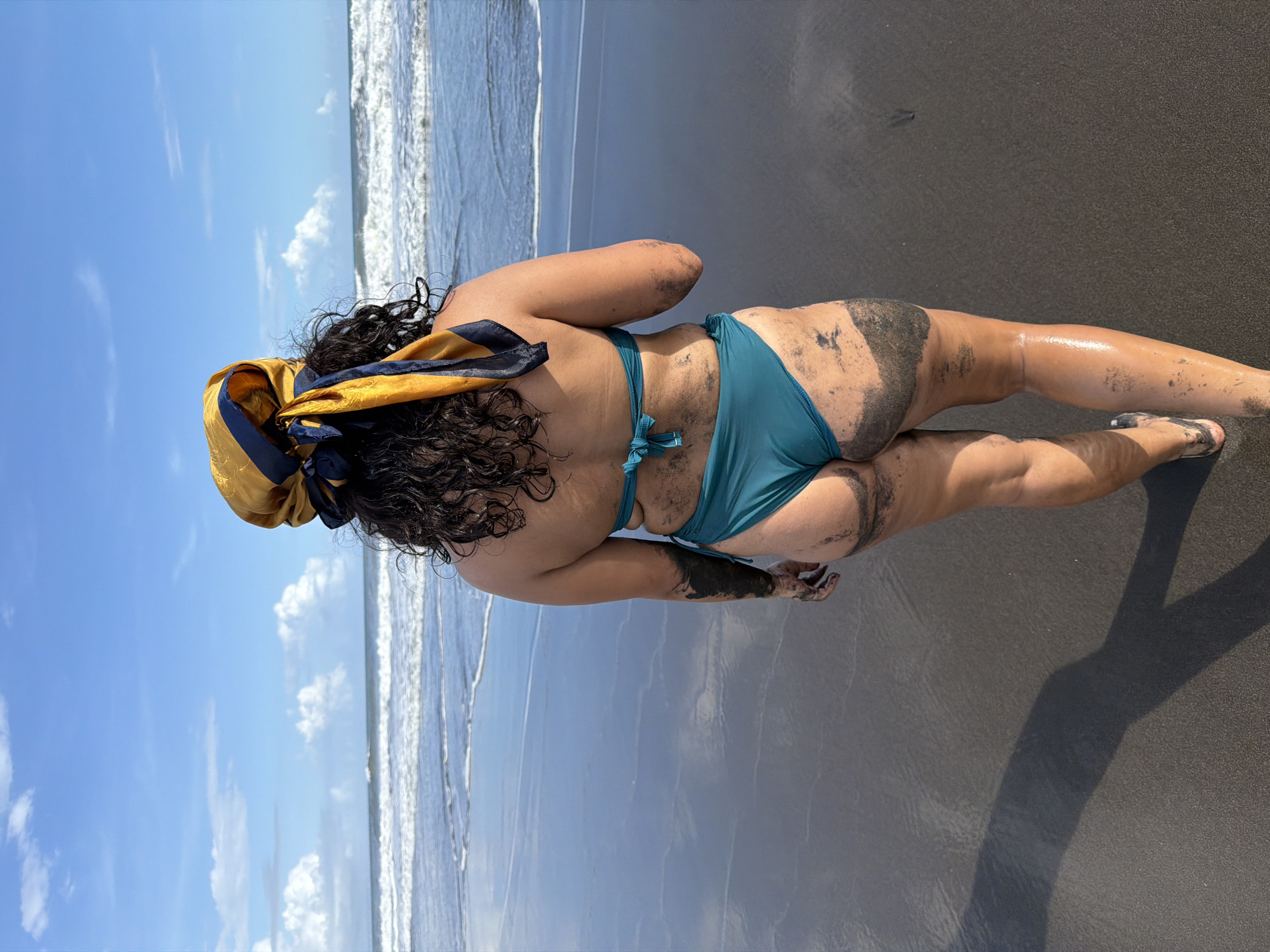 A person is laying on a dark sand beach in Selemadeg Timur, Indonesia. The person, seen from behind, is wearing a teal bikini and a gold and navy blue headscarf. Their dark curly hair is visible beneath the scarf. Parts of their body, including their back, buttocks, arms, and legs, are covered in dark sand. The wet sand of the beach reflects the sky, which is blue with scattered white clouds. Gentle waves are visible in the background.