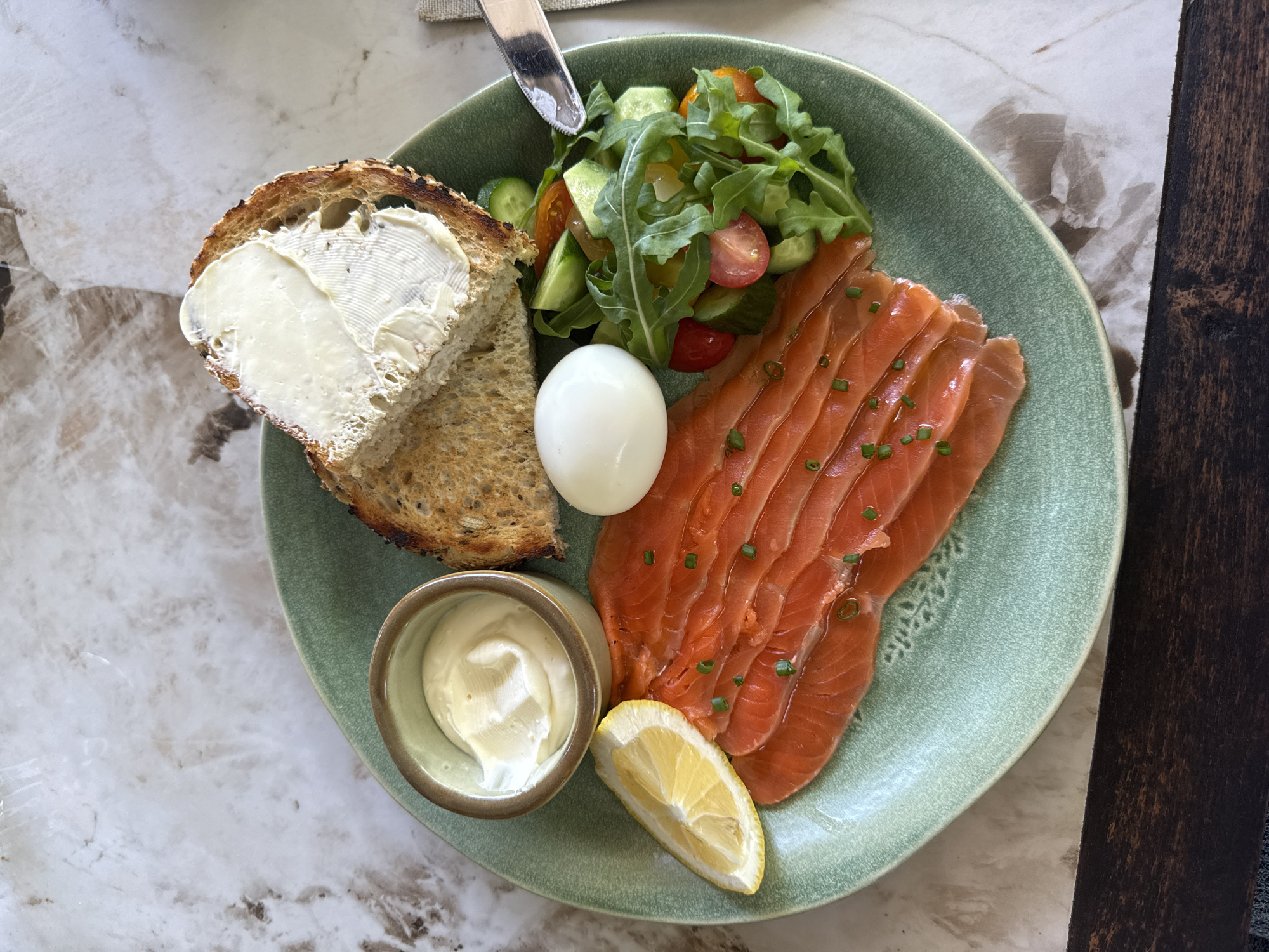 A top-down shot captures a neatly arranged breakfast or brunch plate set on a light-colored, patterned stone table. The main subject is a round, light green ceramic plate with a textured surface, holding a variety of food items.
On the plate, a generous portion of thinly sliced smoked salmon, garnished with chopped green chives, occupies the right side. Beside it, a peeled hard-boiled egg sits prominently. Towards the bottom right, a fresh lemon wedge is placed next to the salmon. In the lower left, a small, two-toned ceramic bowl holds a dollop of creamy white spread, likely cream cheese or a similar condiment.
The left side of the plate features a thick slice of toasted, possibly whole-grain bread, with a visibly crisp crust and a soft, spread-on layer of butter or cream cheese. Above the bread, a vibrant small salad is piled, consisting of green arugula leaves, halved cherry tomatoes, and sliced cucumbers. A silver knife is partially visible at the top, resting on the salad.
The lighting suggests natural daylight, indicating the meal is served during the day. The background includes a dark wooden surface on the right, providing a contrasting texture to the light-colored table and the food-laden plate. There is no visible text in the image. The overall presentation is clean and appealing, showcasing a healthy and balanced meal.