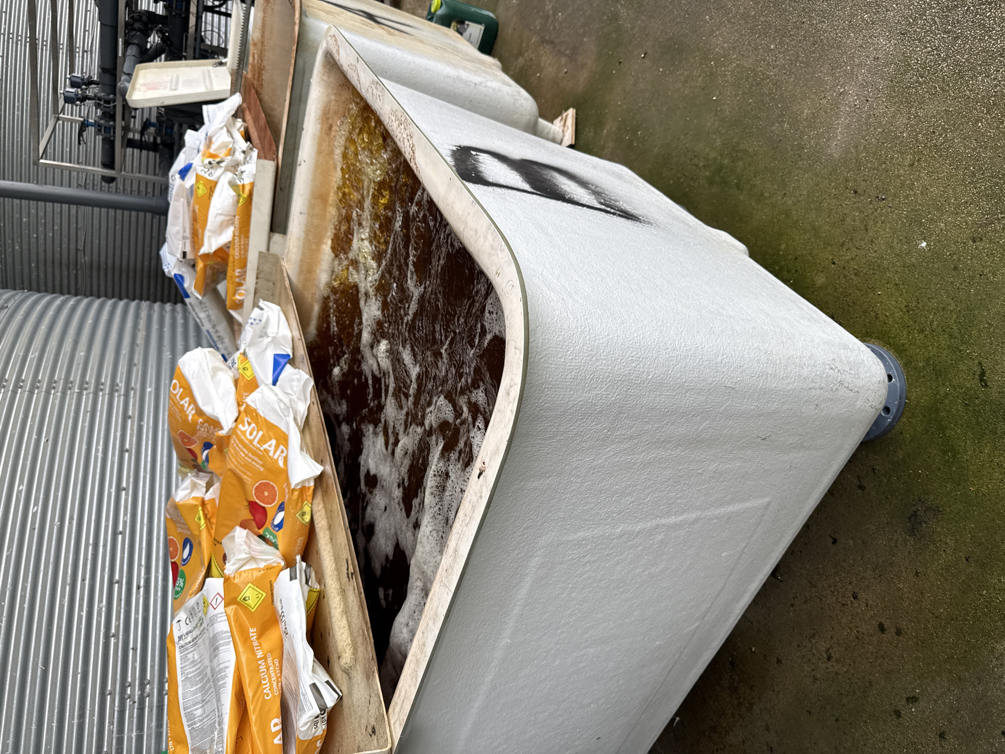 The image shows an outdoor scene with agricultural or horticultural elements. In the foreground, there is a large, white, likely plastic container filled with a dark, foamy liquid. To the left of the container, there is a wooden structure holding multiple bags of fertilizer, specifically "SOLAR Calcium Nitrate". The backdrop includes a corrugated metal structure, possibly a building or a fence, and what appears to be some type of plumbing or irrigation system. The ground is a wet, concrete surface. There is no evidence to confirm that the image was taken in the city of Stara Zagora, Bulgaria. There are no people visible.