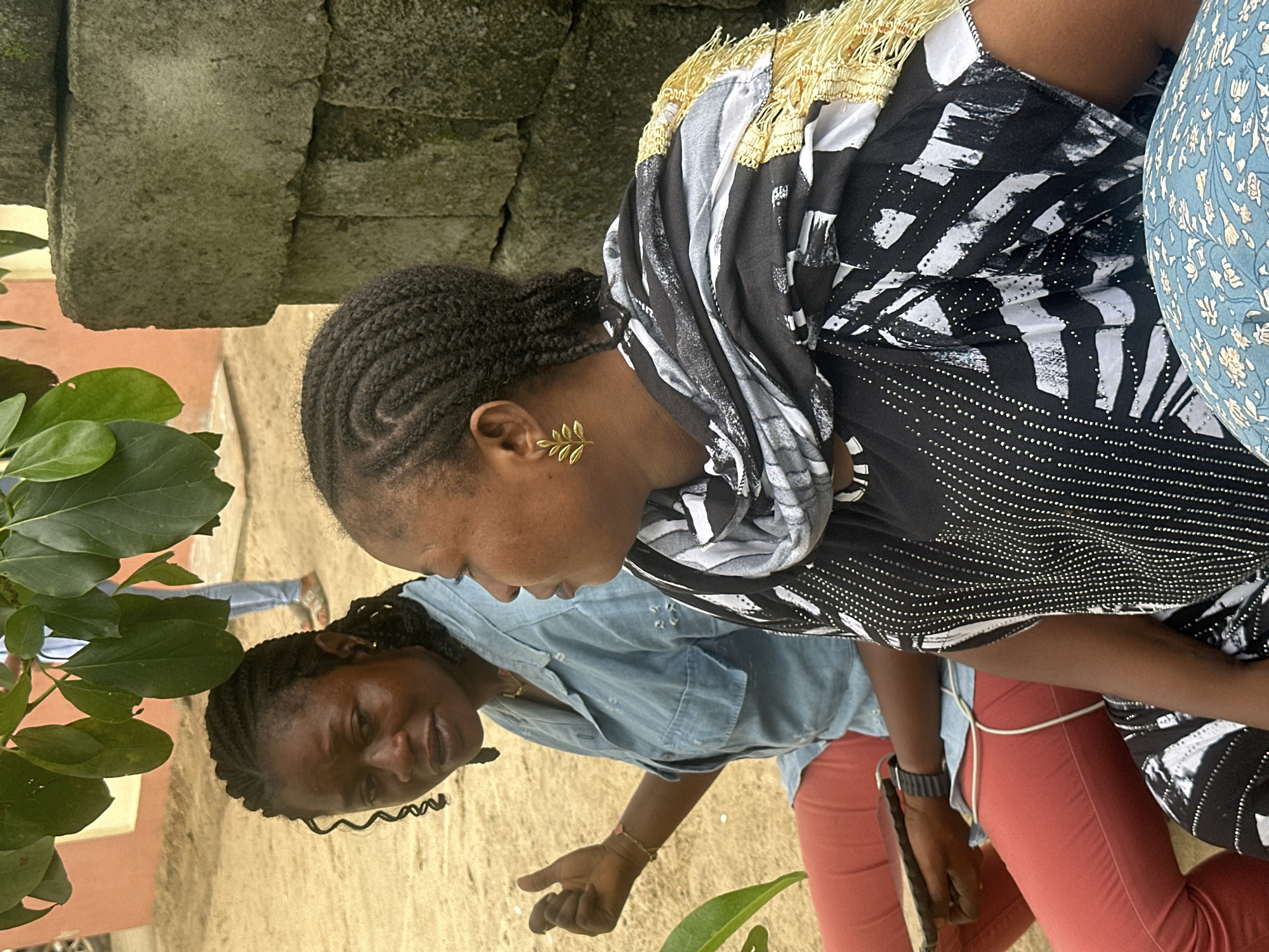 In Ganyingbo, Nigeria, two women are seated outdoors under the shade of tree foliage. The woman on the left, with braided hair and prominent side curls, wears a light blue denim shirt and coral-colored trousers. She holds a dark rectangular object, possibly a phone or tablet, in her left hand, and a thin, light-colored wire is visible connecting her to her lower body. She gestures with her right hand, palm facing outward, fingers slightly curled. The woman on the right, also with braided hair, is adorned with a gold leaf-shaped earring in her left ear and wears a black and white patterned top with a black and white scarf featuring gold fringing around her neck and shoulders. Her posture suggests she is looking to her right. Behind them, a rough stone wall is visible. The ground appears to be sandy soil. A person in the background, partially obscured by leaves, is wearing a blue shirt and light-colored trousers.