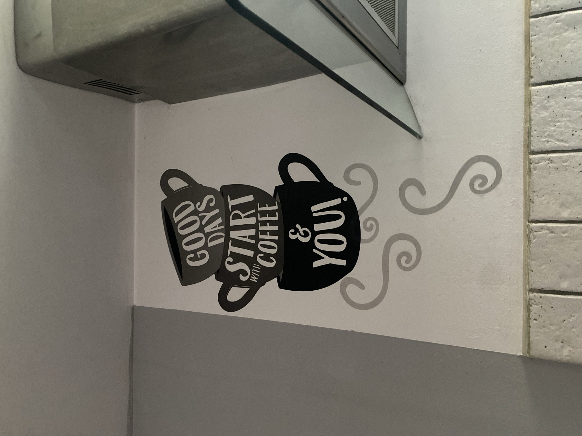 The image shows a wall with a decorative decal featuring three stacked coffee cups. The cups have text that reads "GOOD DAYS START WITH COFFEE & YOU!". The decal is black and white. To the right of the wall, a portion of a kitchen range hood with a glass canopy is visible. The wall appears to be painted white and is situated in a domestic setting, likely a kitchen. There are no people or animals present in the image. The image was taken in Wrocław, Poland.