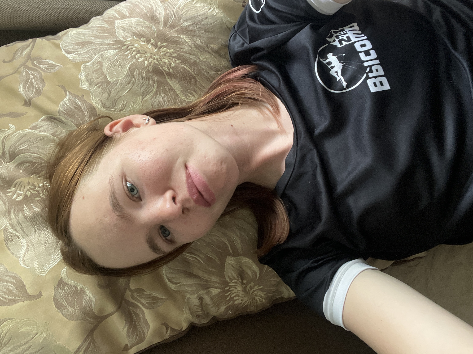 A young Caucasian woman with light brown hair and blue eyes is lying on a pillow. She is wearing a black t-shirt with a white logo on it, which appears to depict a climber. The pillow she is lying on has a floral pattern in beige and gold. The background is a couch with a dark brown textured fabric. The image appears to be a selfie taken from a high angle. There is no discernible location within the city of Chaikovsky, Russia.